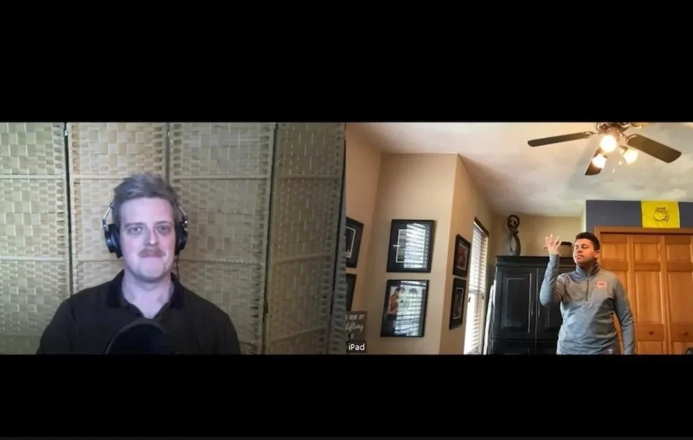 A split-screen image showing two men in a virtual meeting. The man on the left has light brown hair, a mustache, wearing headphones and a black shirt, against a textured, beige background. The man on the right has dark hair, wearing a gray jacket, standing in a room with framed pictures on the wall, a ceiling fan, and wooden cabinets, raising his right hand.