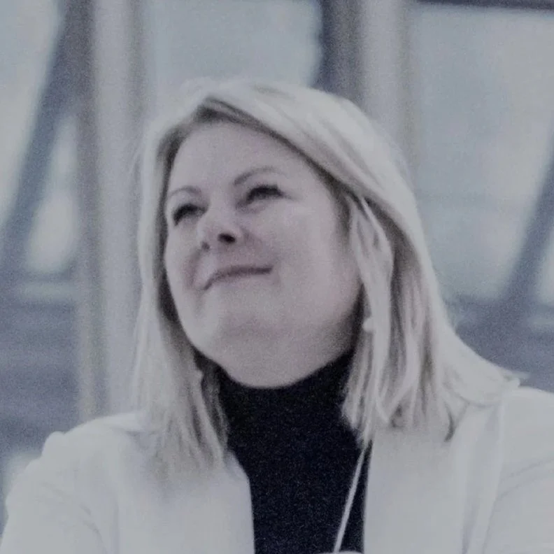 A woman with blonde hair smiling, wearing a black turtleneck and white blazer, in an indoor setting.