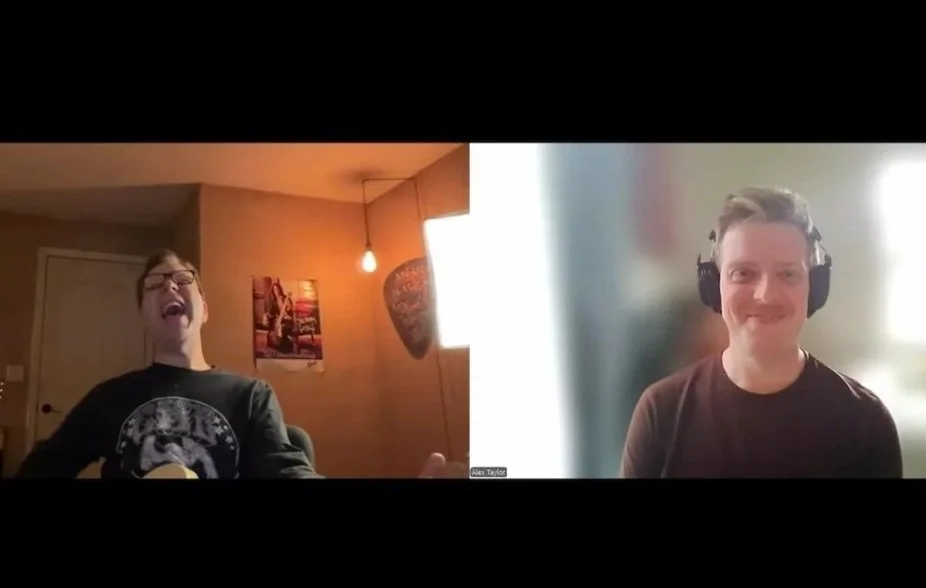 A video call showing two men, one on the left wearing glasses and a black t-shirt with a lion graphic, laughing with his mouth open, and the other on the right wearing headphones and a black t-shirt, smiling. The background on the left is warm with a hanging light and a wall painting, while the background on the right is blurred and bright.
