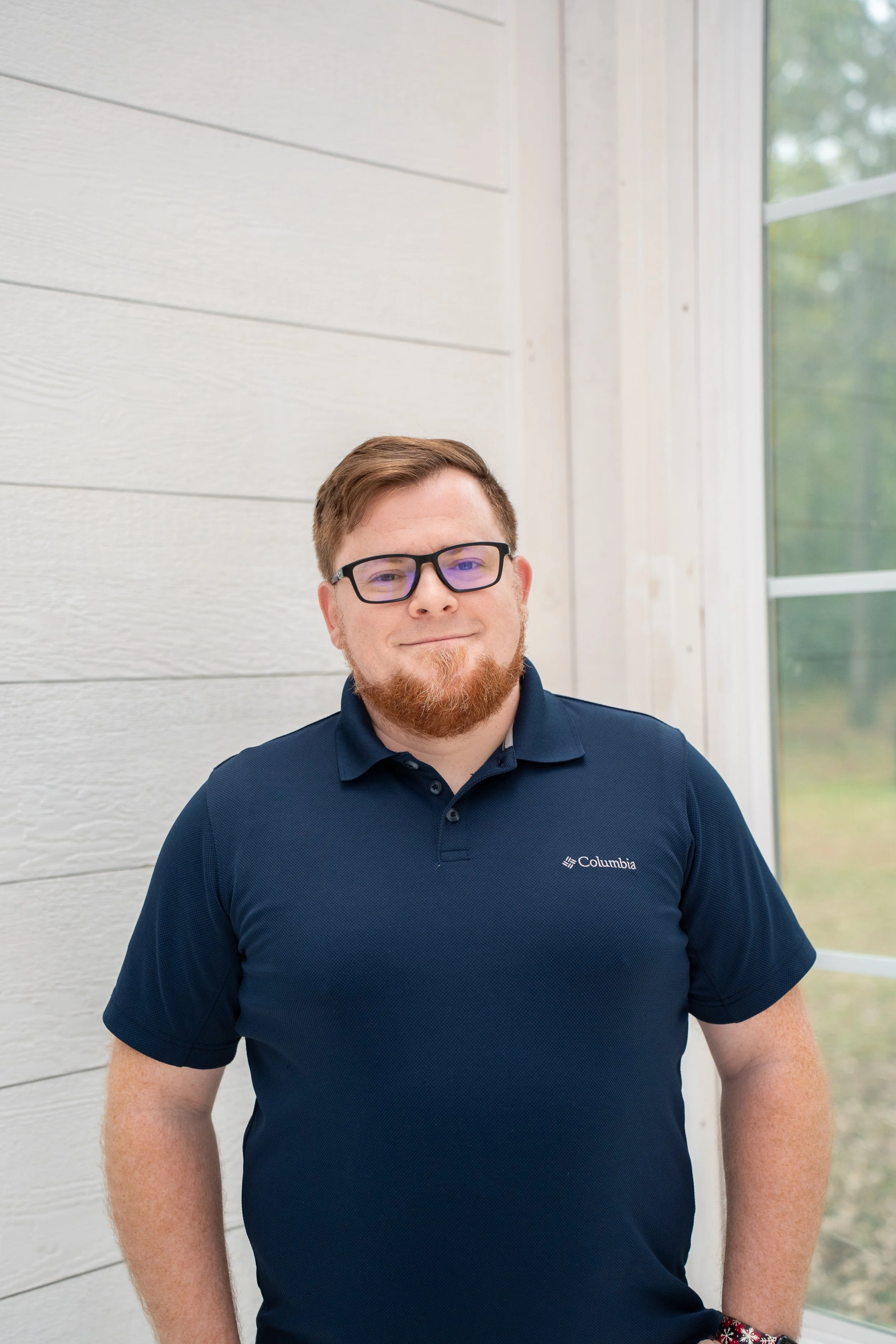 A man with glasses and a beard standing indoors near a window, wearing a navy blue Columbia polo shirt.