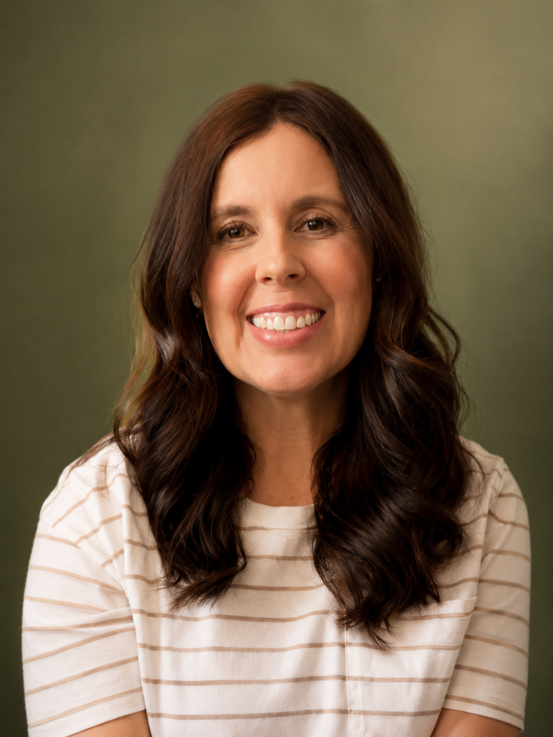 A woman with long, wavy dark brown hair smiling at the camera, wearing a cream and beige striped shirt, against a green background.