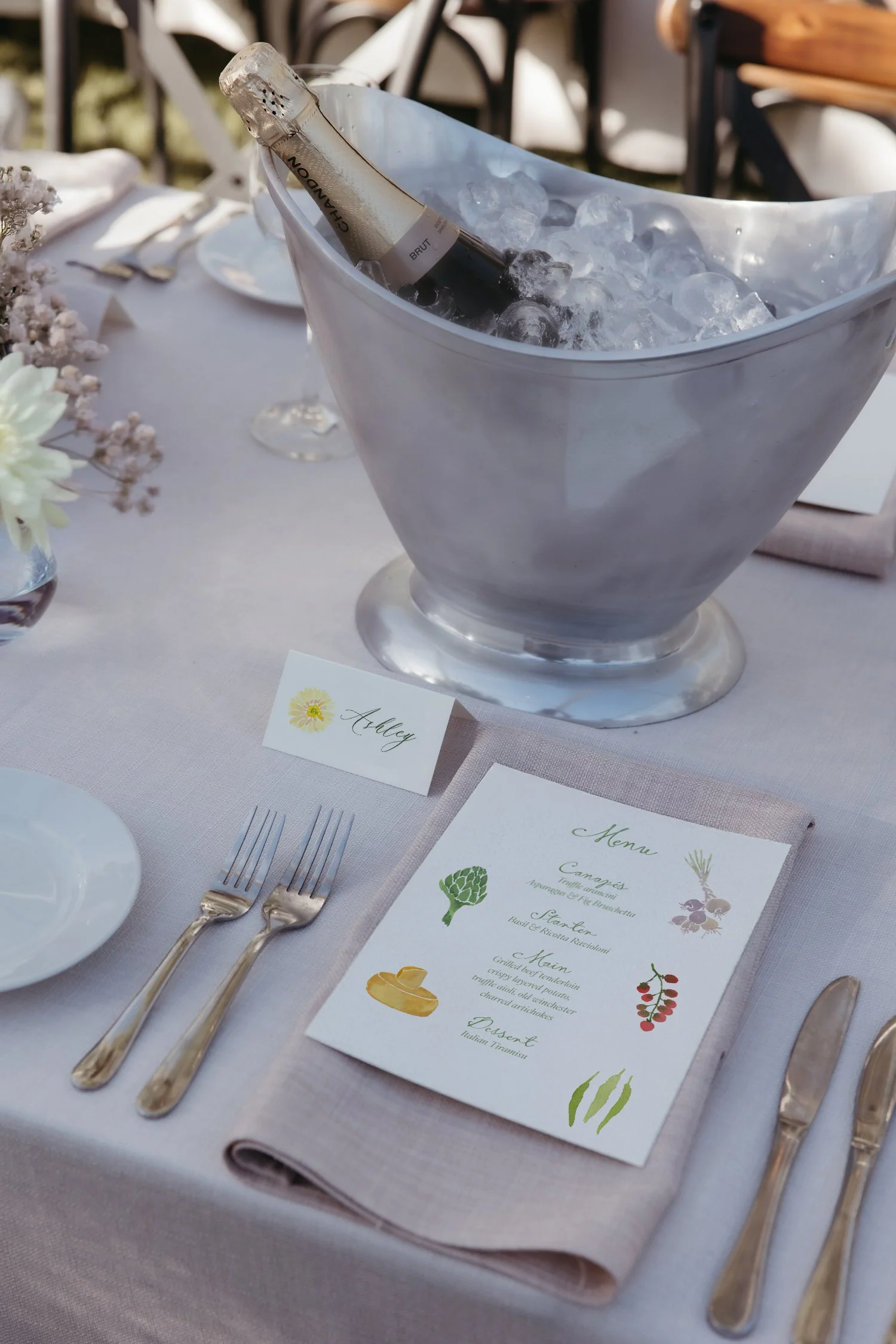 A table setting with a wine bucket filled with ice and a bottle of sparkling wine, a menu, a name card labeled 'Aubrey,' and silverware, at an outdoor event or wedding reception.
