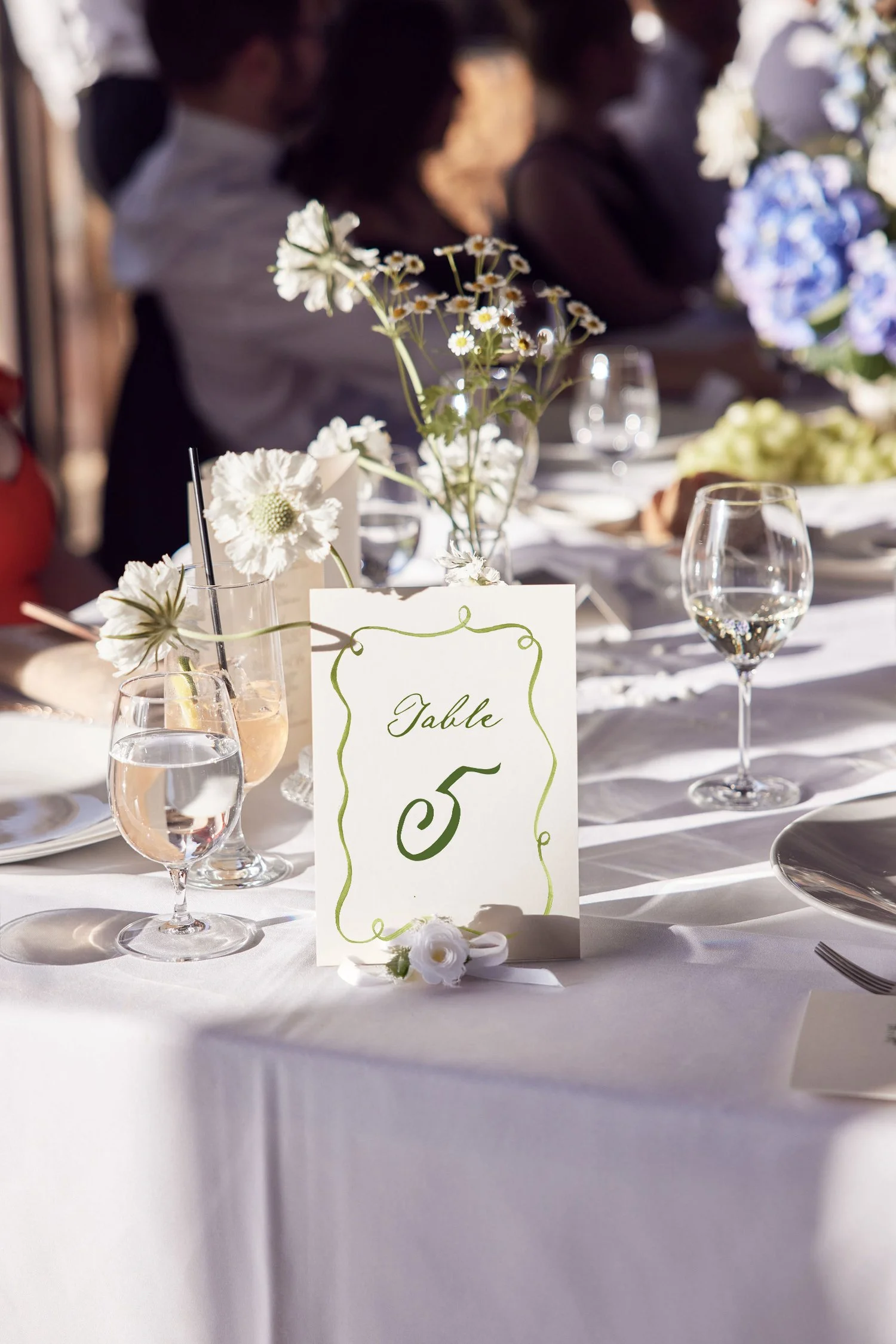 A table setting at a wedding or elegant event, featuring a white tablecloth, floral centerpieces with white and purple flowers, wine glasses, a table number sign labeled 'Table 5' in green cursive, and sunlight casting shadows.