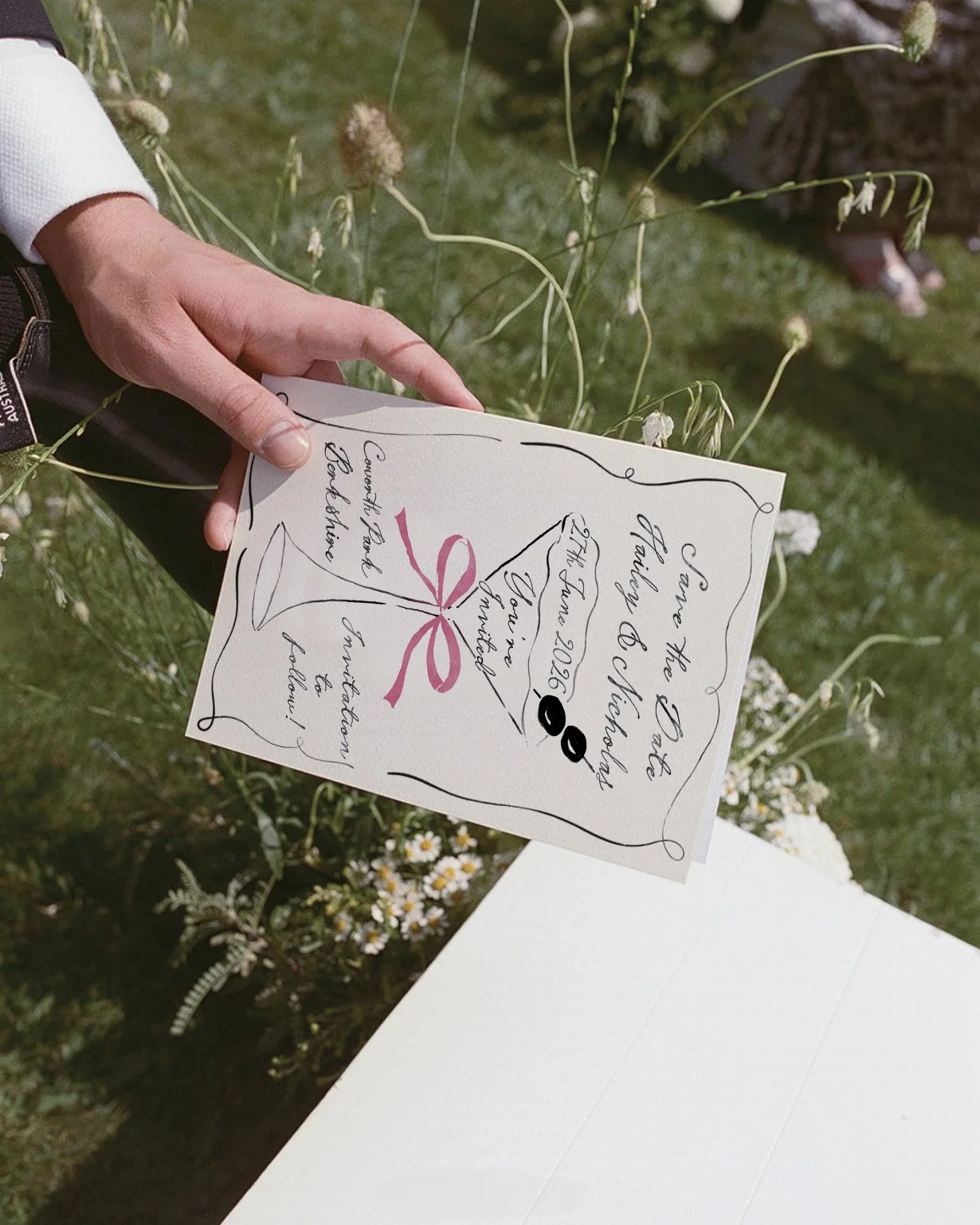 A person holding a wedding invitation with black and pink handwritten script, outside among greenery and small white daisy flowers.