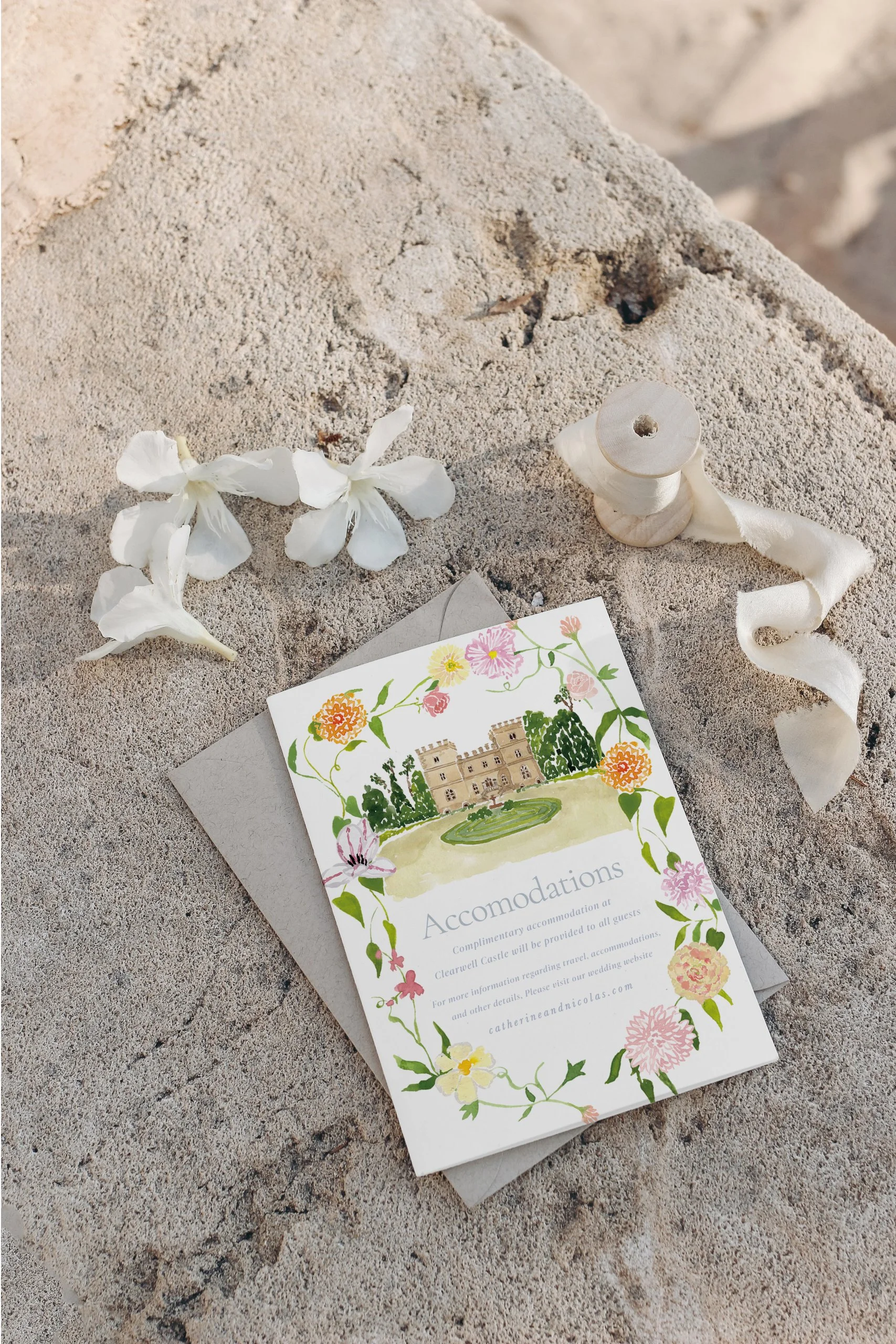 A wedding or event card with floral border, illustration of a castle, and the word 'Accommodations' on sand with white flowers, a spool of ribbon, and a linen envelope nearby.
