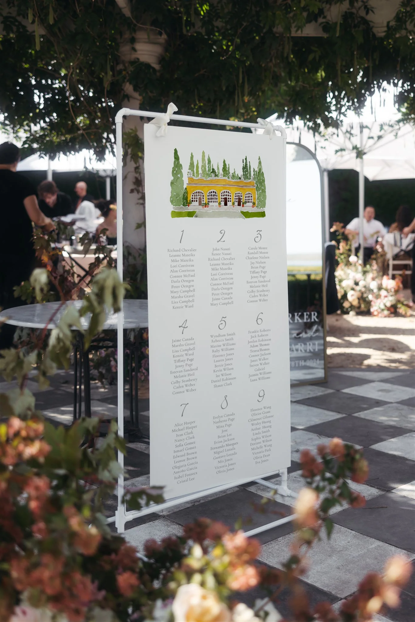 A white standing sign with a list of names, an illustrated building at the top, surrounded by greenery, set outdoors with people dining and flowers in the background.