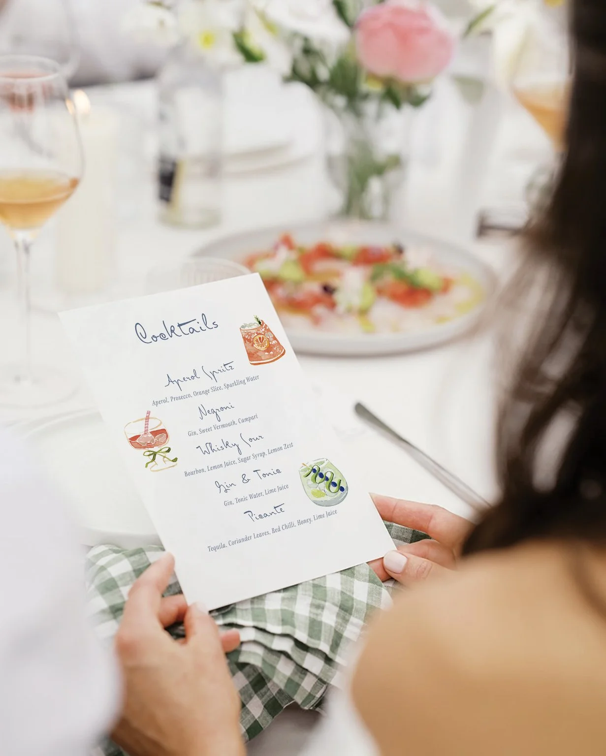 Person holding a printed cocktail menu at a table with glasses of wine, a plate of food, and a floral arrangement in the background.