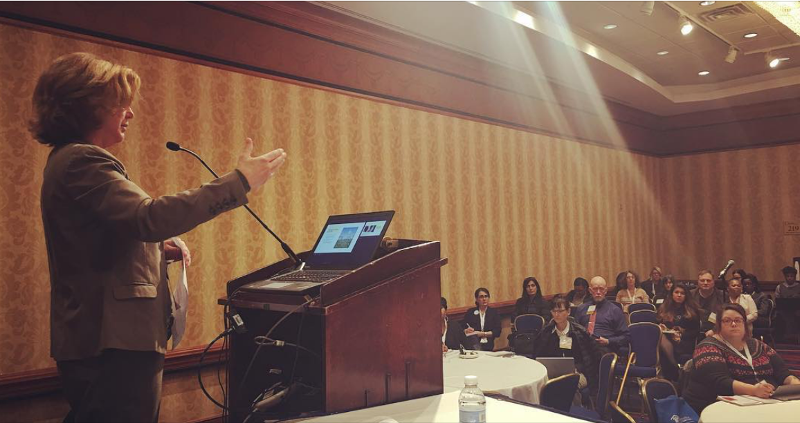 A woman giving a presentation at a conference, standing behind a wooden podium with a laptop, addressing an audience seated in a theater-style room.