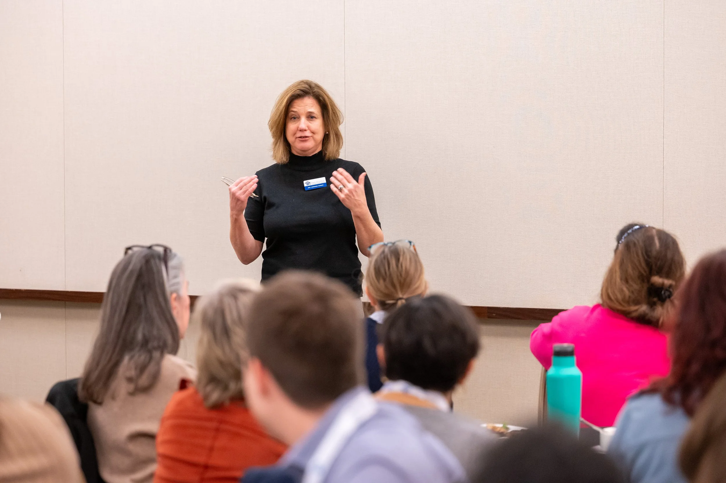 A woman speaking to a seated audience in a conference room. She is wearing a black top and gesturing with her hands, with a name tag on her chest. The audience is diverse and appears attentive.