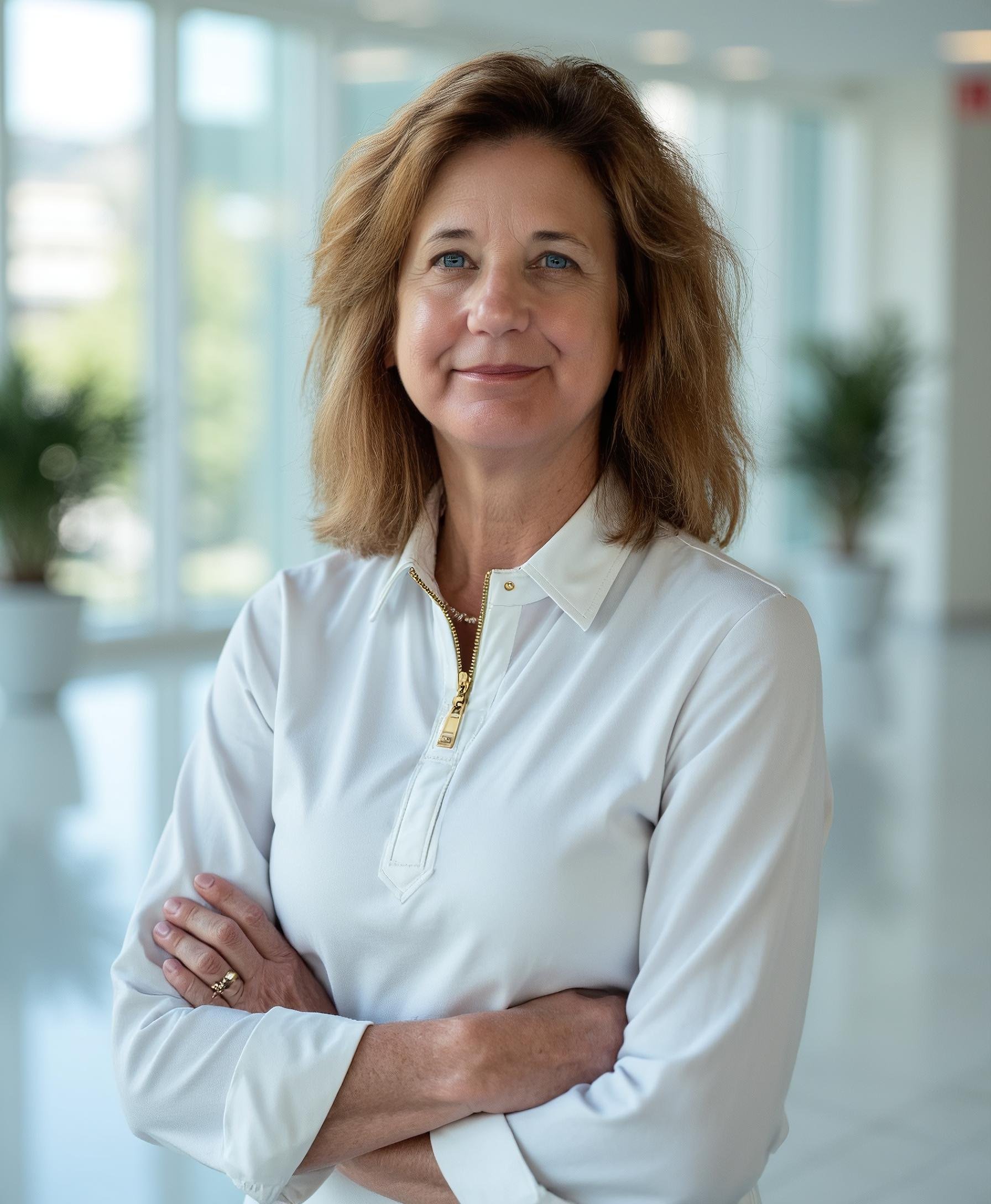 A middle-aged woman with shoulder-length light brown hair and blue eyes standing with her arms crossed in a bright, modern indoor space with large windows and potted plants in the background.