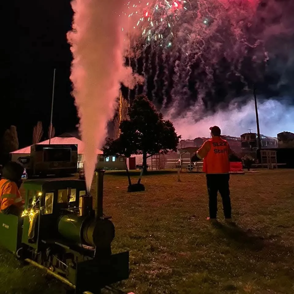 A miniature steam train blows steam at a railway event in the North of England, with fireworks in the sky at a night time event with a celebration of railway heritage