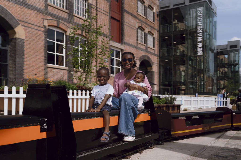 Miniature train ride with people, featuring a family sitting on a miniature train carriage, with a historic industrial styled building in the background, a white picket fence and some newly planted trees, an educational experience.