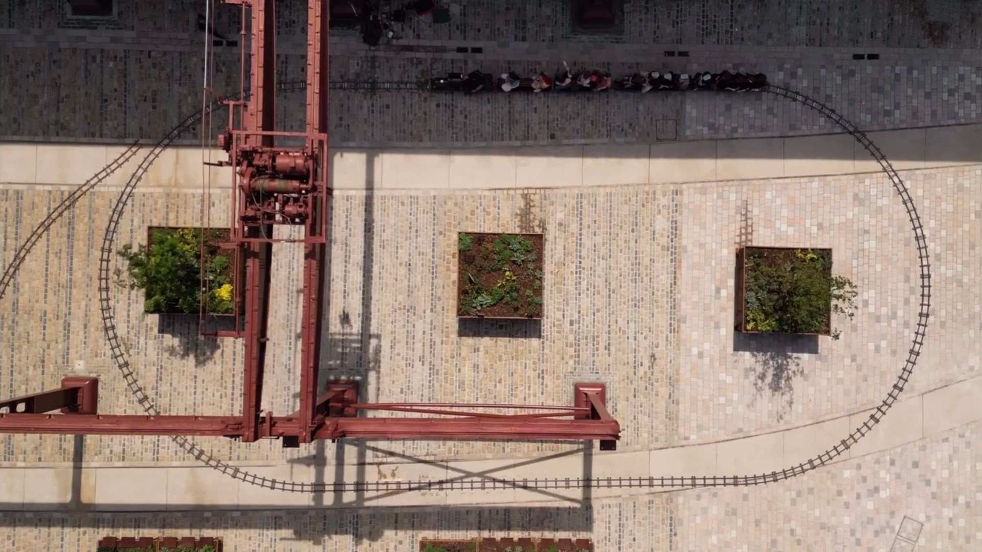 Arial view of oval train track laid out on stone courtyard with three planter boxes and a restored vintage overhead crane structure casting a shadow across the miniature steam train which can be seen on part of the oval