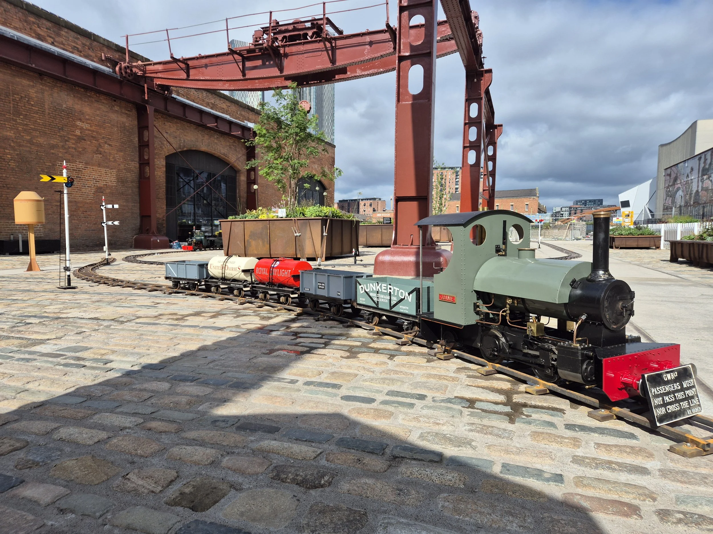 A small vintage train with green and red colors runs on railway tracks in an urban area, with brick buildings, a tree, and modern high-rise buildings in the background. The train is on display in a museum as part of a school holiday event program.