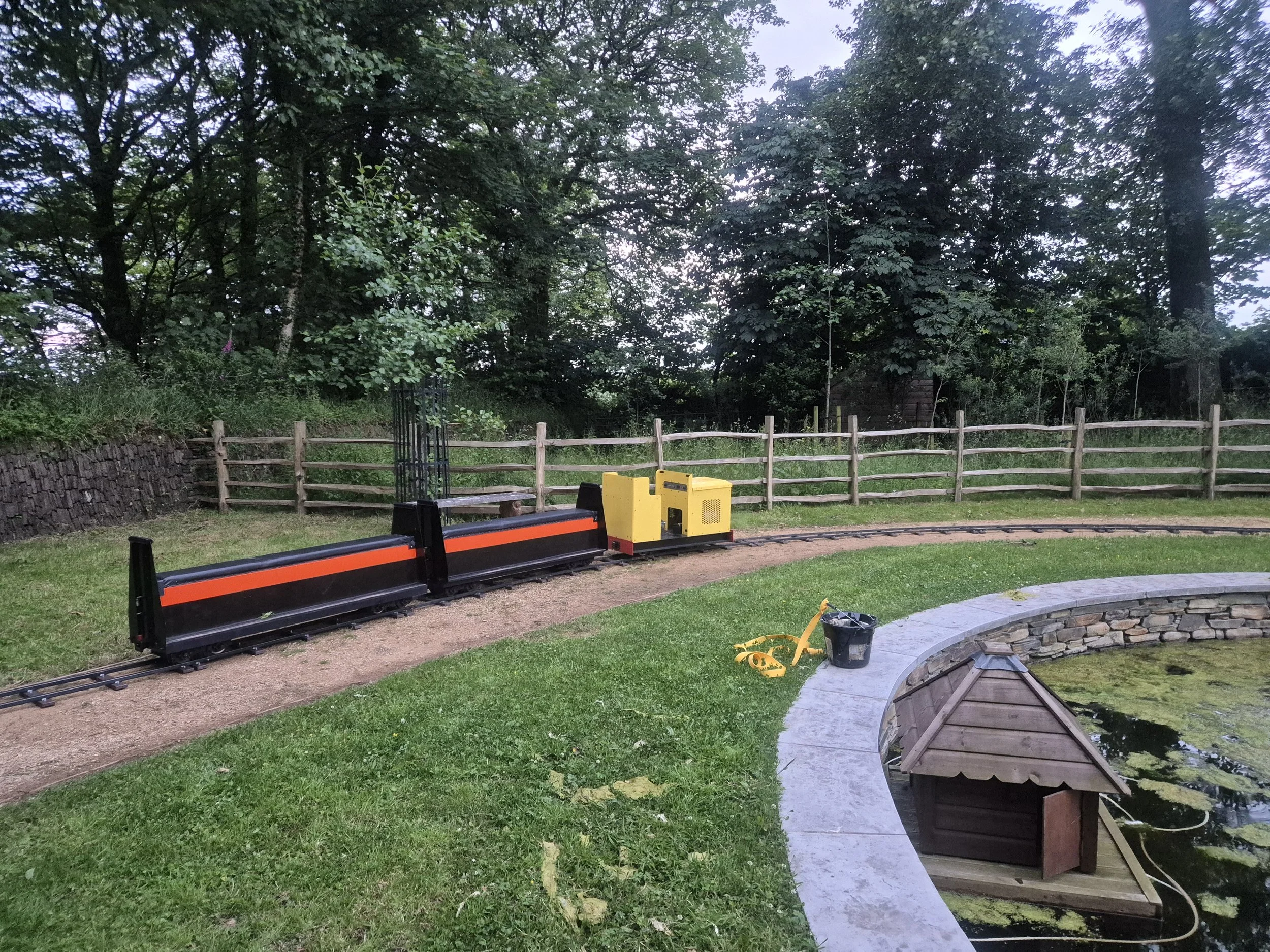A yellow miniature railway locomotive waits with two carriages in a leafy green setting for passengers to board and take a journey on the railway at a specialist event hire