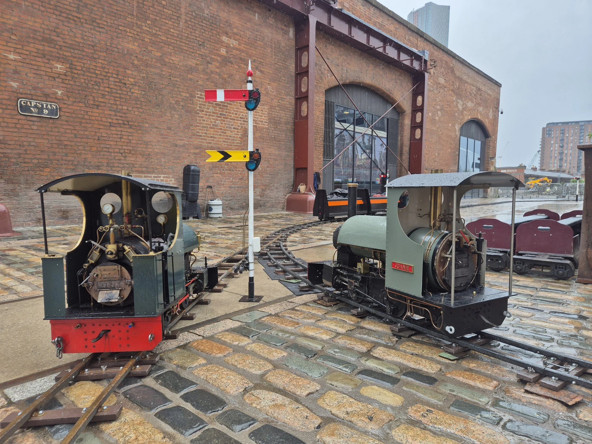 Miniature steam train locomotives on cobblestone street near a brick building with a large window and railroad crossing signals.