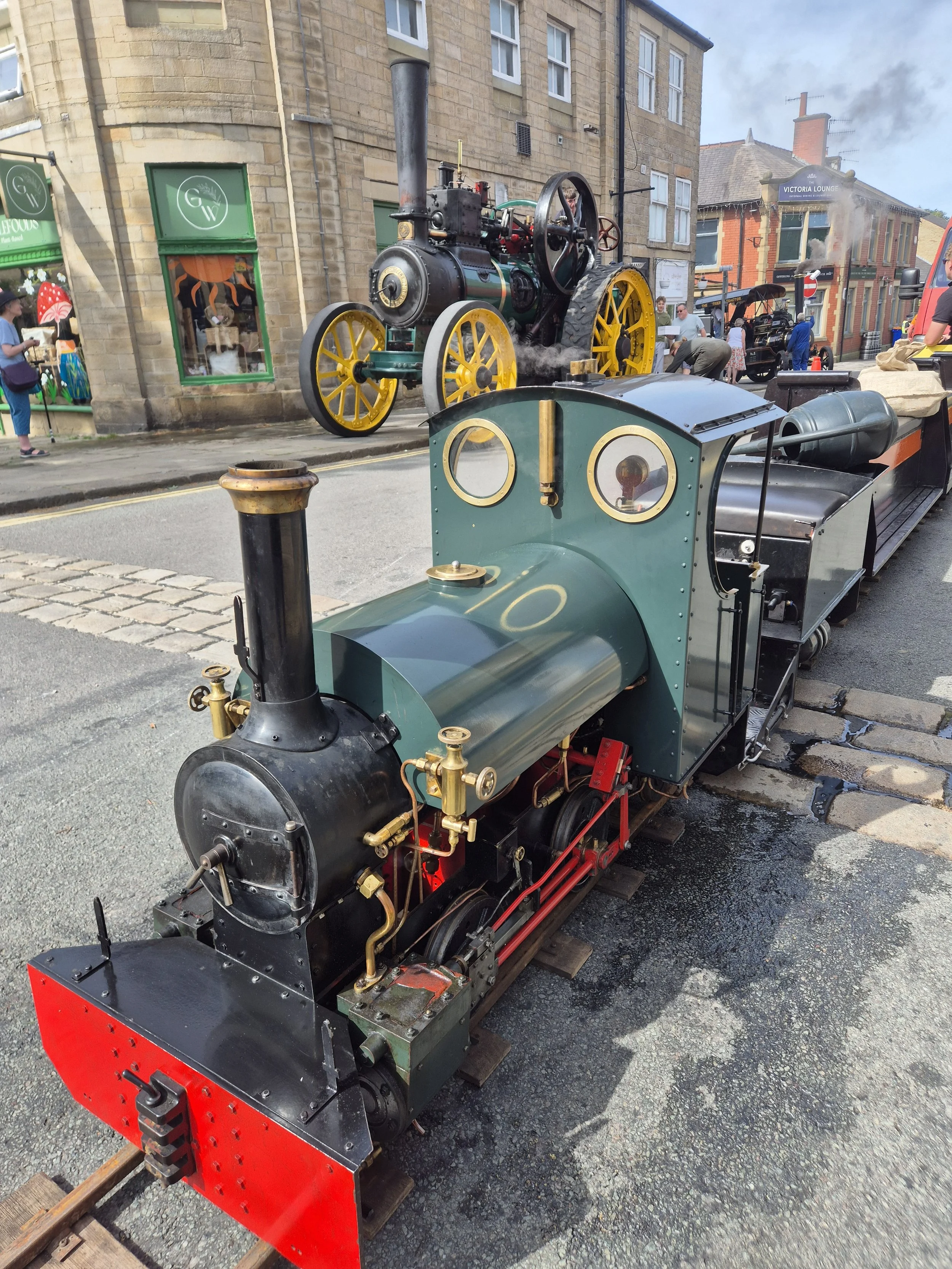A vintage miniature steam locomotive displayed outdoors on tracks, with people observing and a building with storefronts in the background.