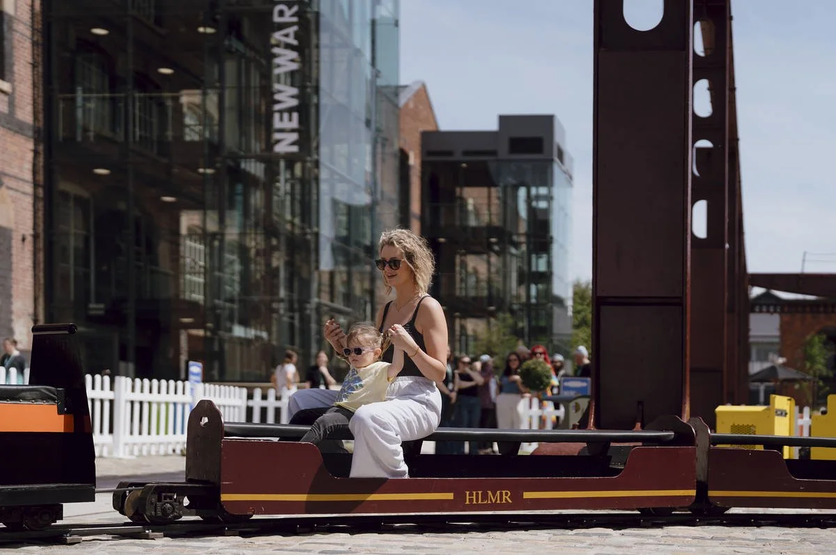 A woman and a young girl sitting on a miniature train ride in an outdoor urban setting, with buildings and people visible in the background. Part of an attraction at a museum in England.