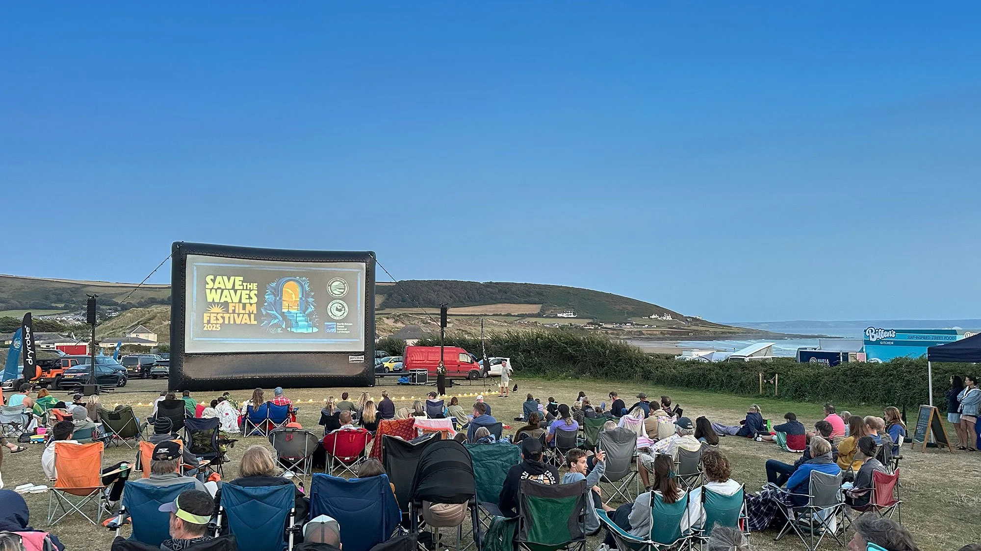Photo from a field in Croyde, overlooking the beach with lots of people sat watching a large outdoor screen which shows the title 'Save The Waves Film Festival 2025'