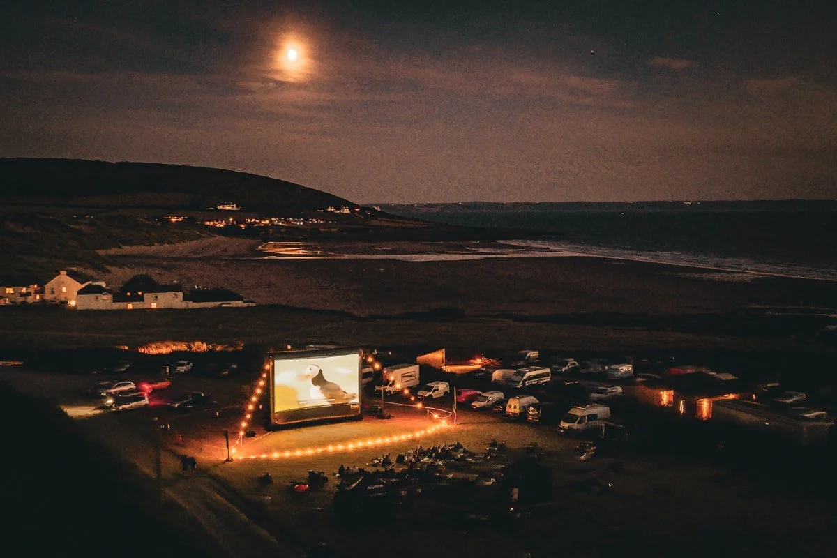 Atmospheric photo of the festival after sunset, showing lights around the big screen, which features an image of a puffin, with Croyde Bay in the background.