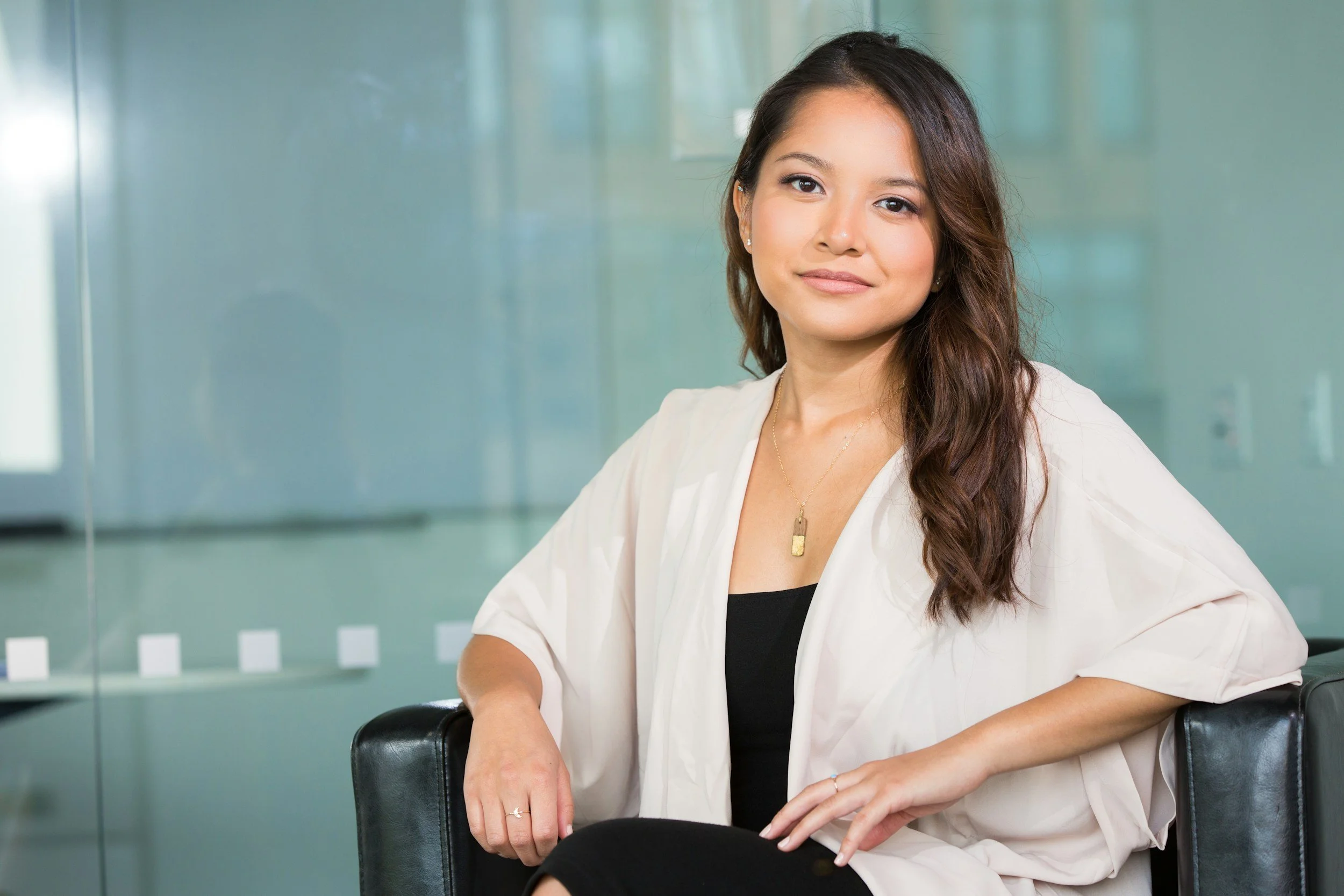 A woman with long brown hair, wearing a white blazer over a black top, sitting on a black chair in a modern office setting with a glass wall.
