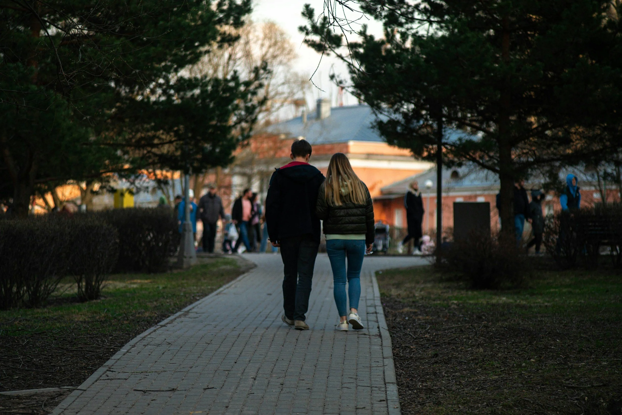 A couple walking hand in hand along a paved pathway in a park, surrounded by trees and other people in the background near a brick building.