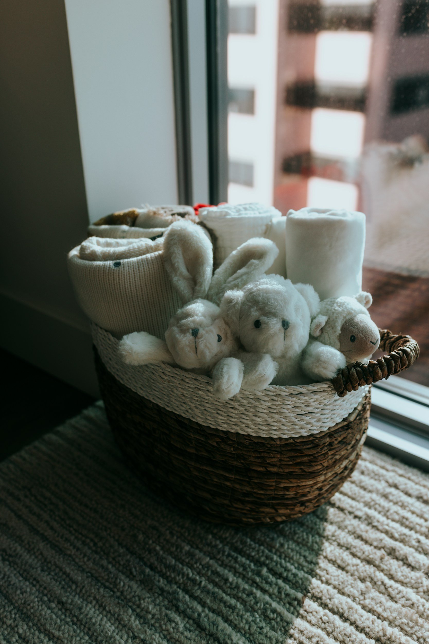 A woven basket filled with plush white teddy bears, rolled white towels, and baby socks, placed on a striped rug near a glass door.