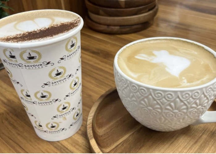 A to-go coffee cup with a hot latte and a white ceramic mug with a frothy cappuccino on a wooden table.