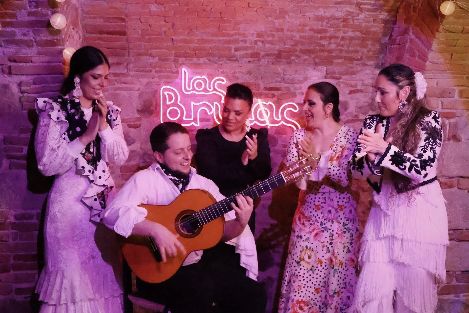 Tres mujeres bailando flamenco con vestimenta tradicional, en un escenario con iluminación rosa y negra.