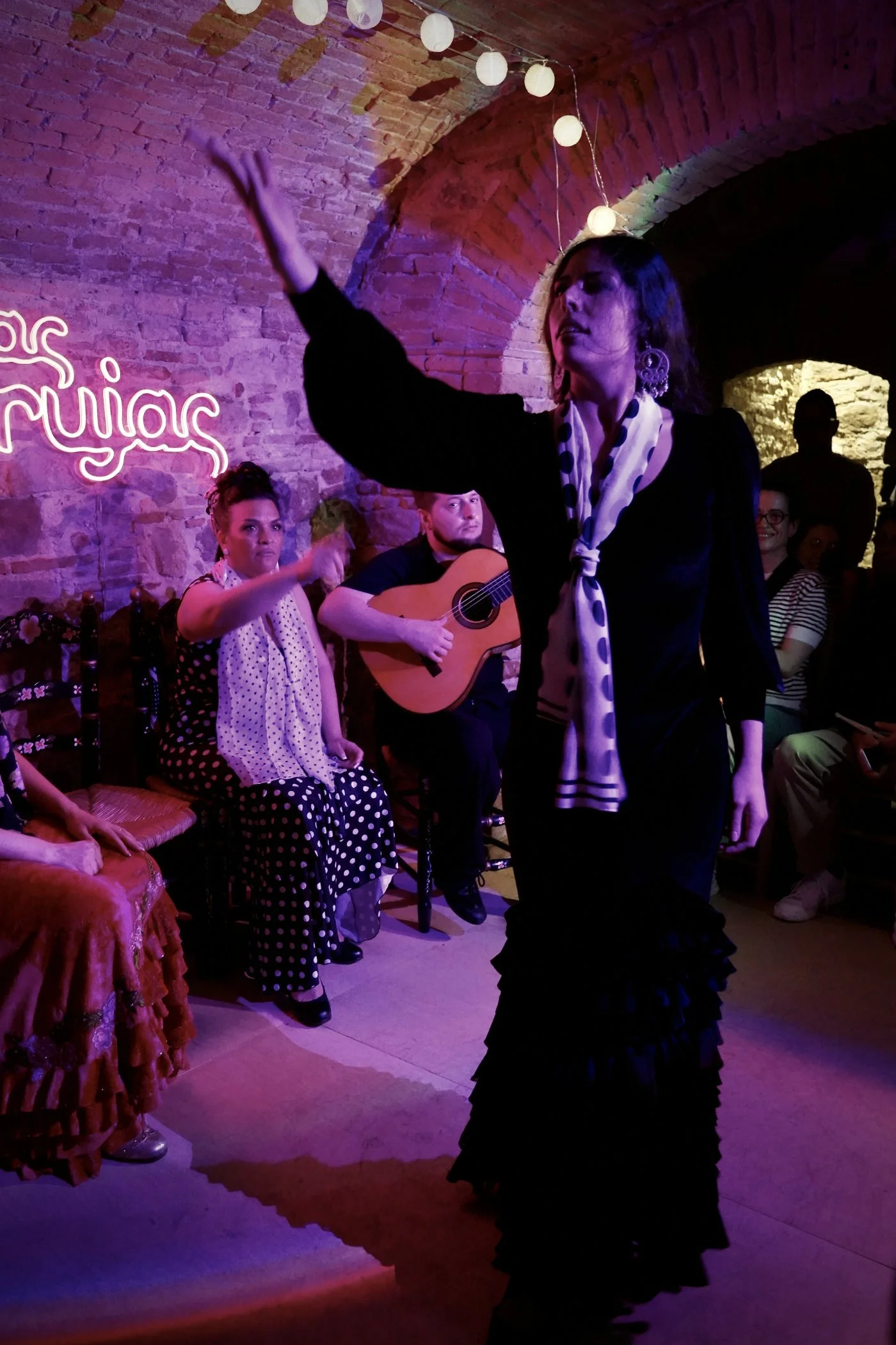 Una mujer en un vestido largo de flamenco, bailando en un ambiente con paredes de ladrillo a la vista y luces de papel en el techo, rodeada de personas, algunas tocando guitarra y otras observando y sonriendo.