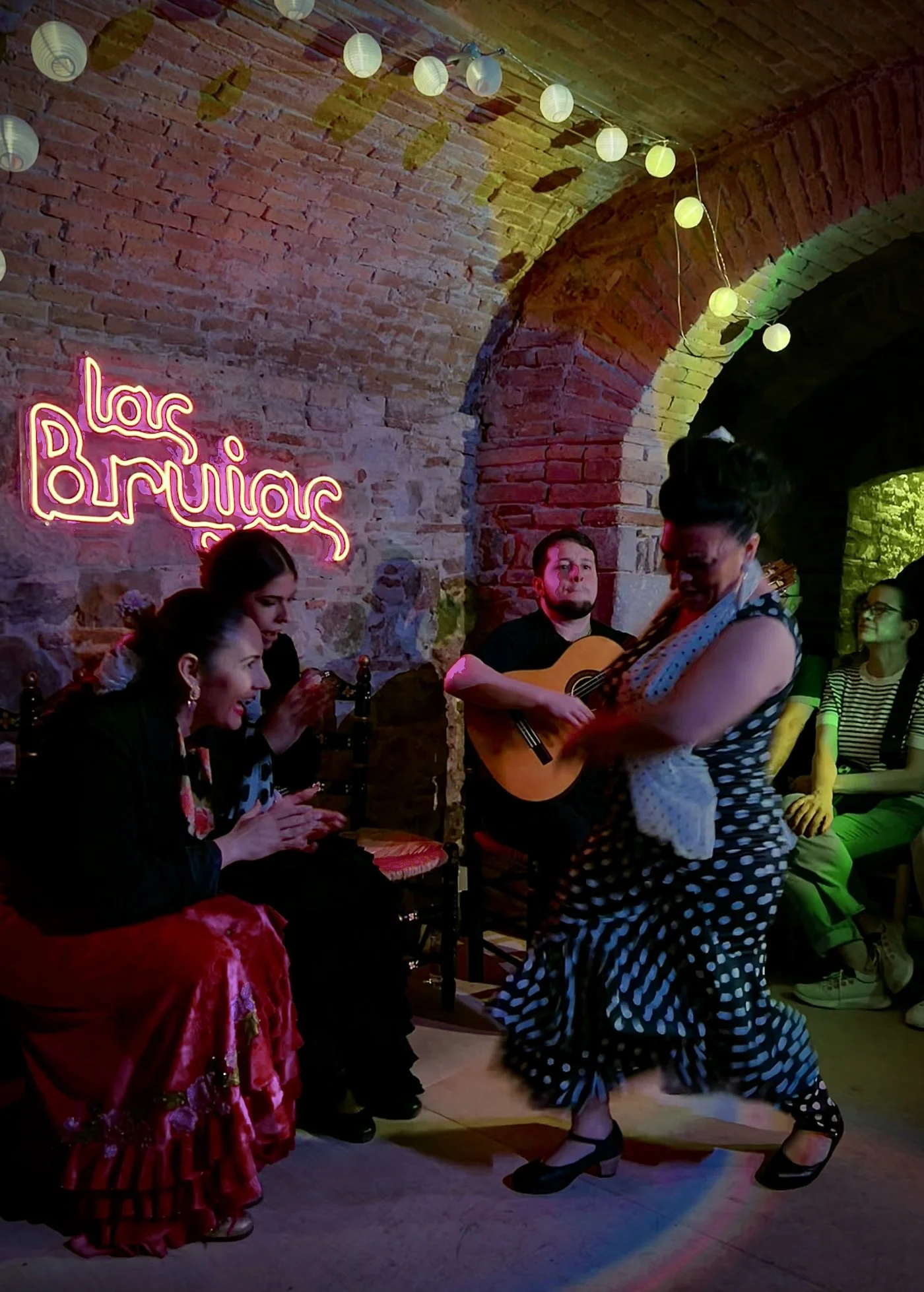 Personas bailando flamenco en un ambiente con paredes de ladrillo y luces de estilo papel. Una mujer con vestido de flamenco negro con lunares blancos y zapatos de tacón baila con una mujer con vestido rosa y lunares negros. En el fondo, un hombre toca la guitarra y hay un cartel de neón que dice 'las brújias'.