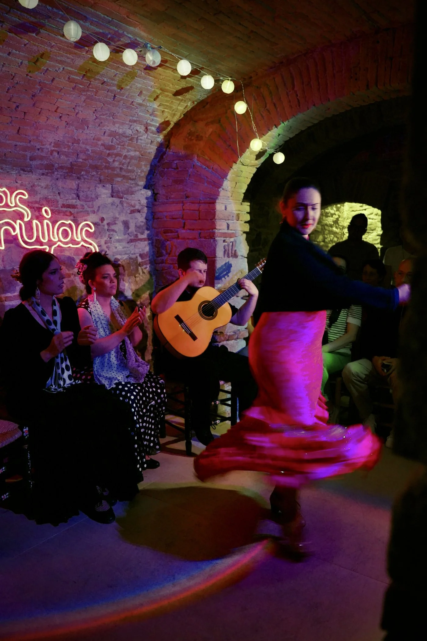 Un grupo de personas sentadas en un lugar con paredes de ladrillo a la vista, decorado con luces de papel y un letrero de neón que dice 'Flamenco'. En el centro, una bailarina vestida con un vestido largo de flamenco rosa y con movimiento, aparece en el acto de bailar, mientras varias personas la observan y una toca la guitarra.