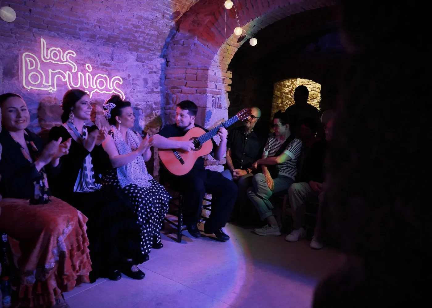 Grupo de personas en un tablao llamado 'Las Brujas', con decoraciones de neón y paredes de ladrillo, disfrutando de una presentación de flamenco en vivo, donde un hombre toca la guitarra y las demás personas aplauden y observan.