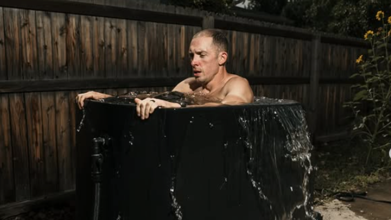 A man is submerged in a black bathtub filled with water outside in a backyard with a wooden fence and some yellow flowers.