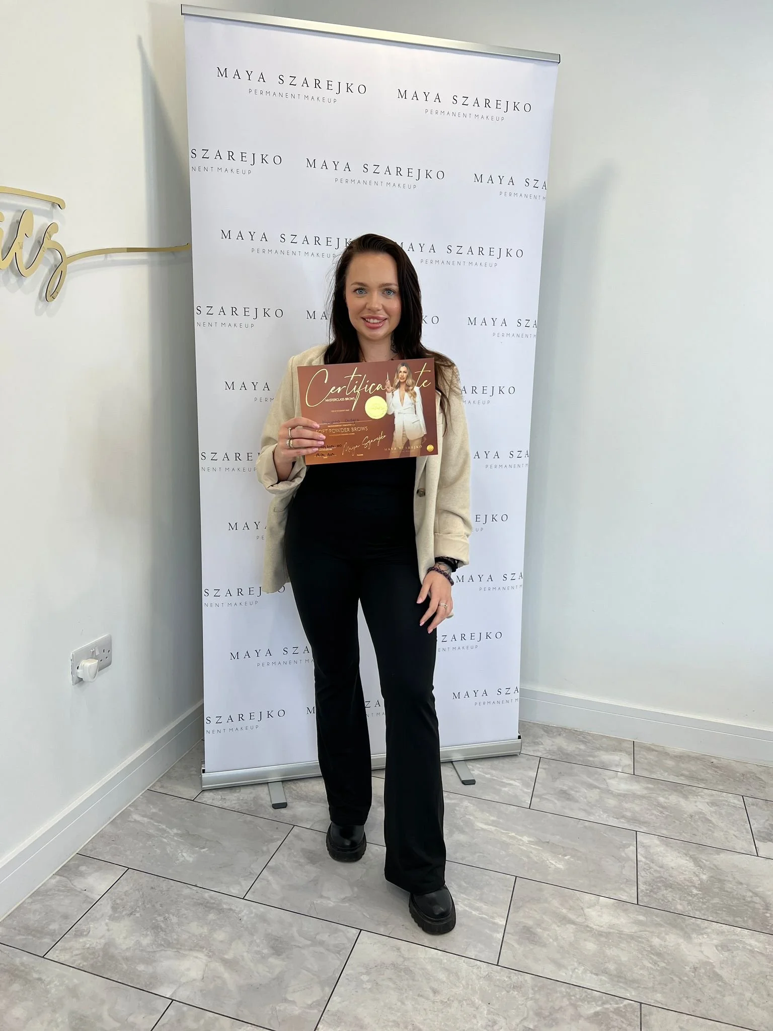 Person holding a certificate in front of a Maya Szarejko Permanent Makeup branded banner.