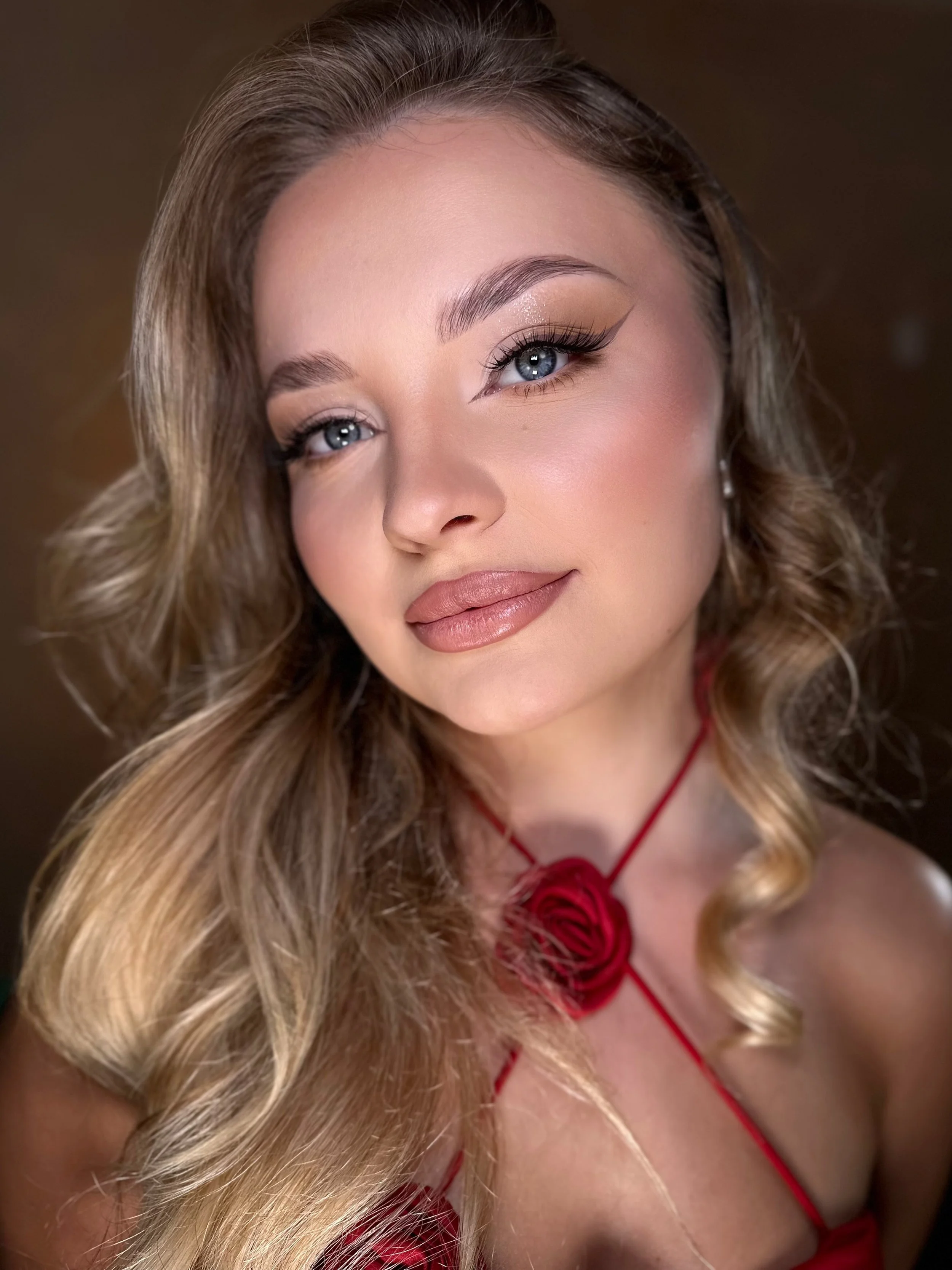 A woman with long blond hair, wearing a red dress with a floral design, and makeup with winged eyeliner and neutral lipstick.