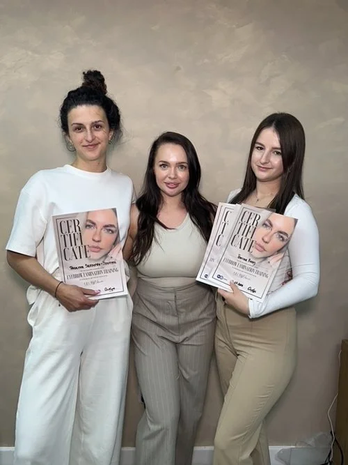 Three women standing against a beige wall, with two on the sides holding certificates. The woman in the center is not holding anything.