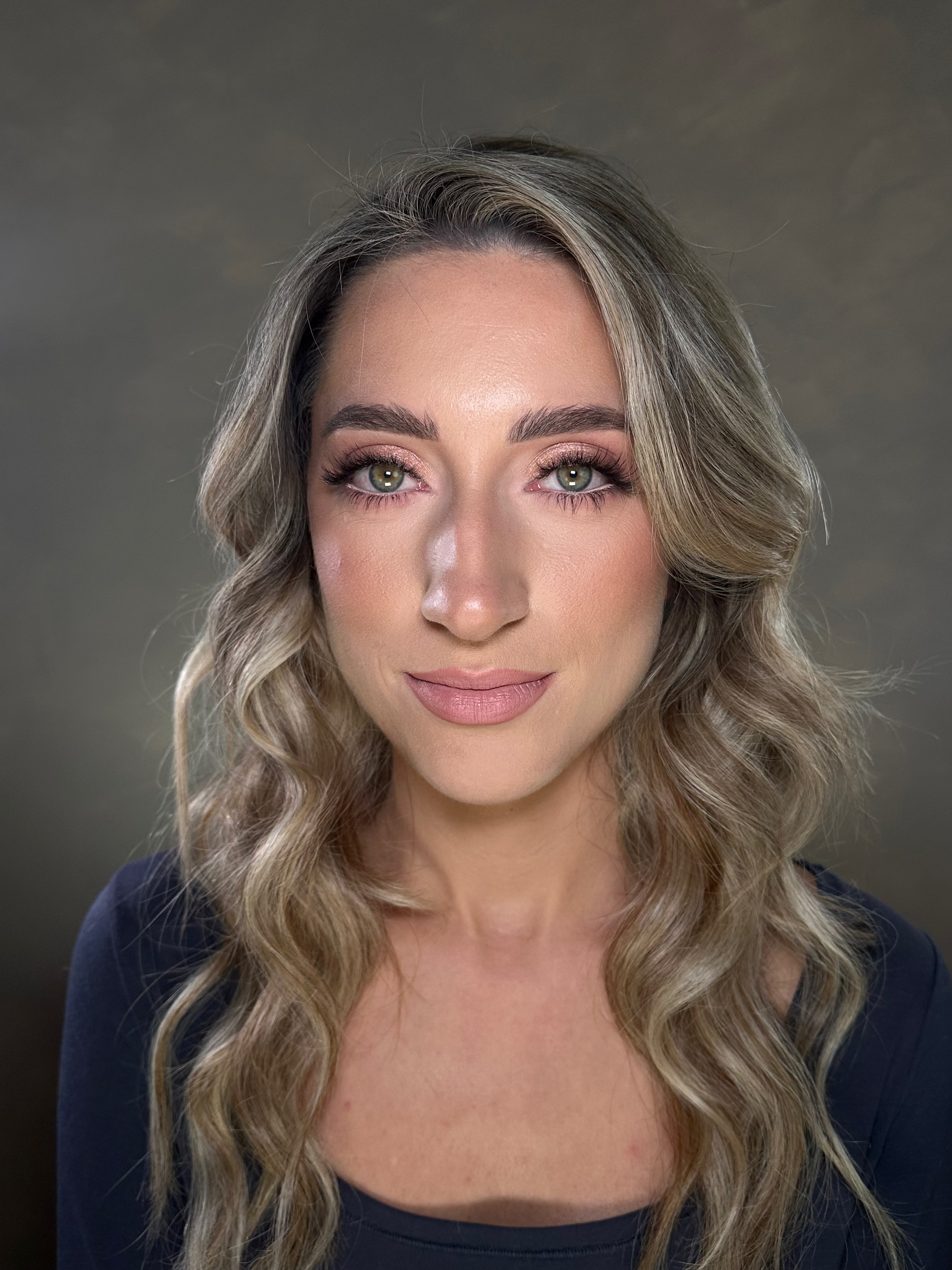 A woman with wavy blond hair and makeup, wearing a black top, looking at the camera against a neutral background.