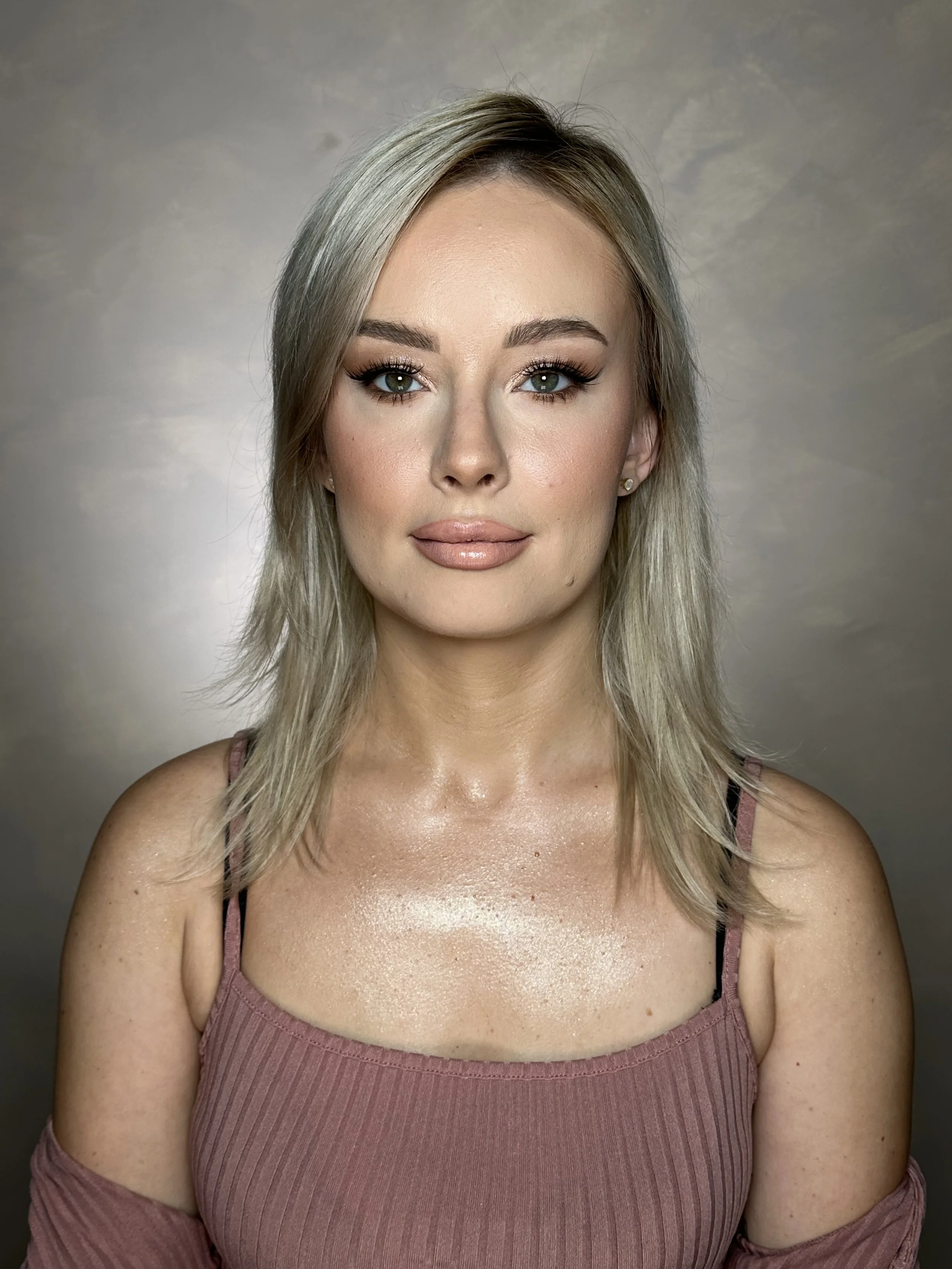 A young woman with blonde hair, wearing makeup, a pink sleeveless top, and black bra straps showing, looking directly at the camera with a neutral expression against a neutral background.