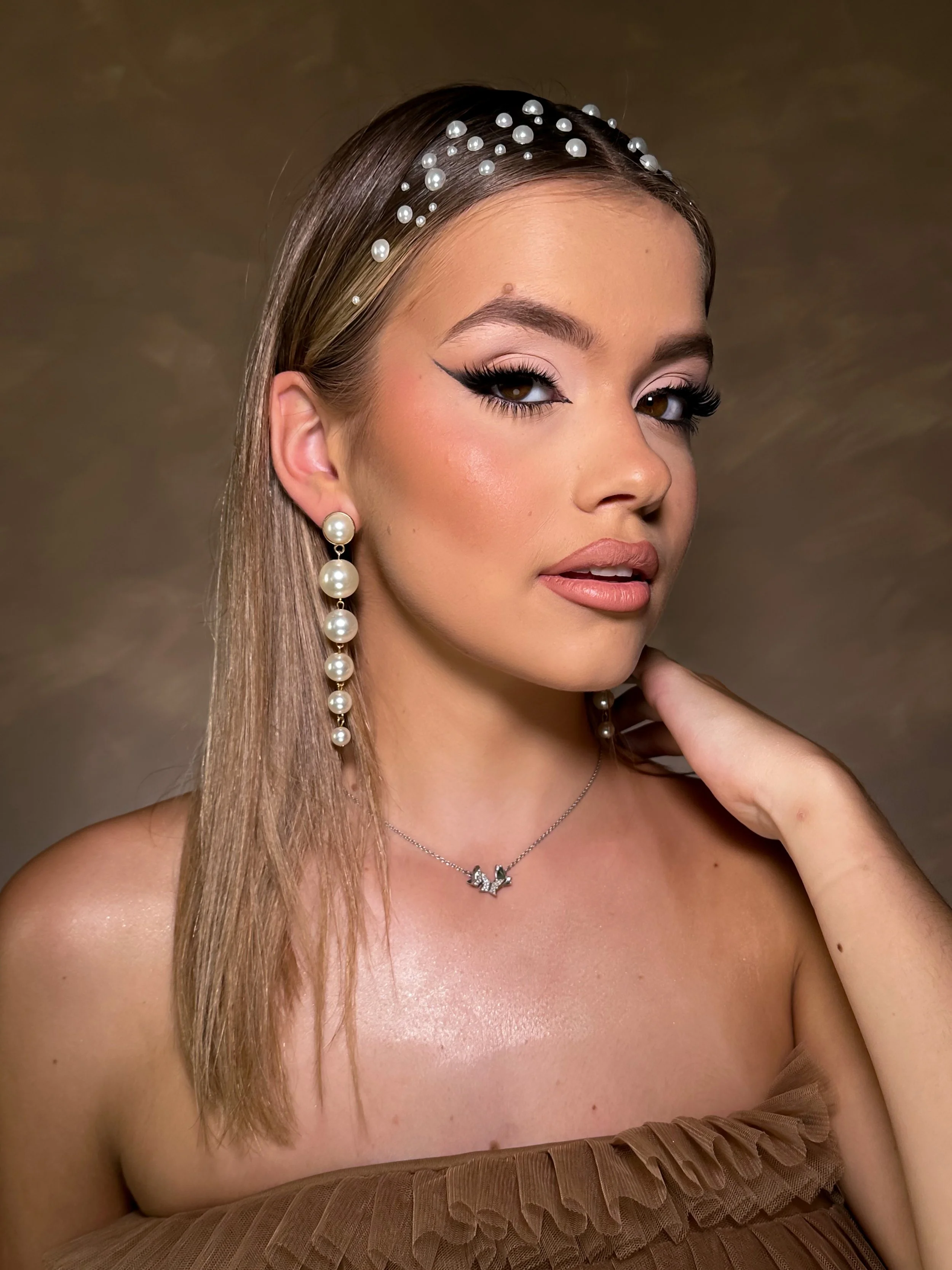 A woman with pearl hair accessories, pearl earrings, and a necklace, wearing makeup and a brown strapless dress with ruffles, poses against a gold background.