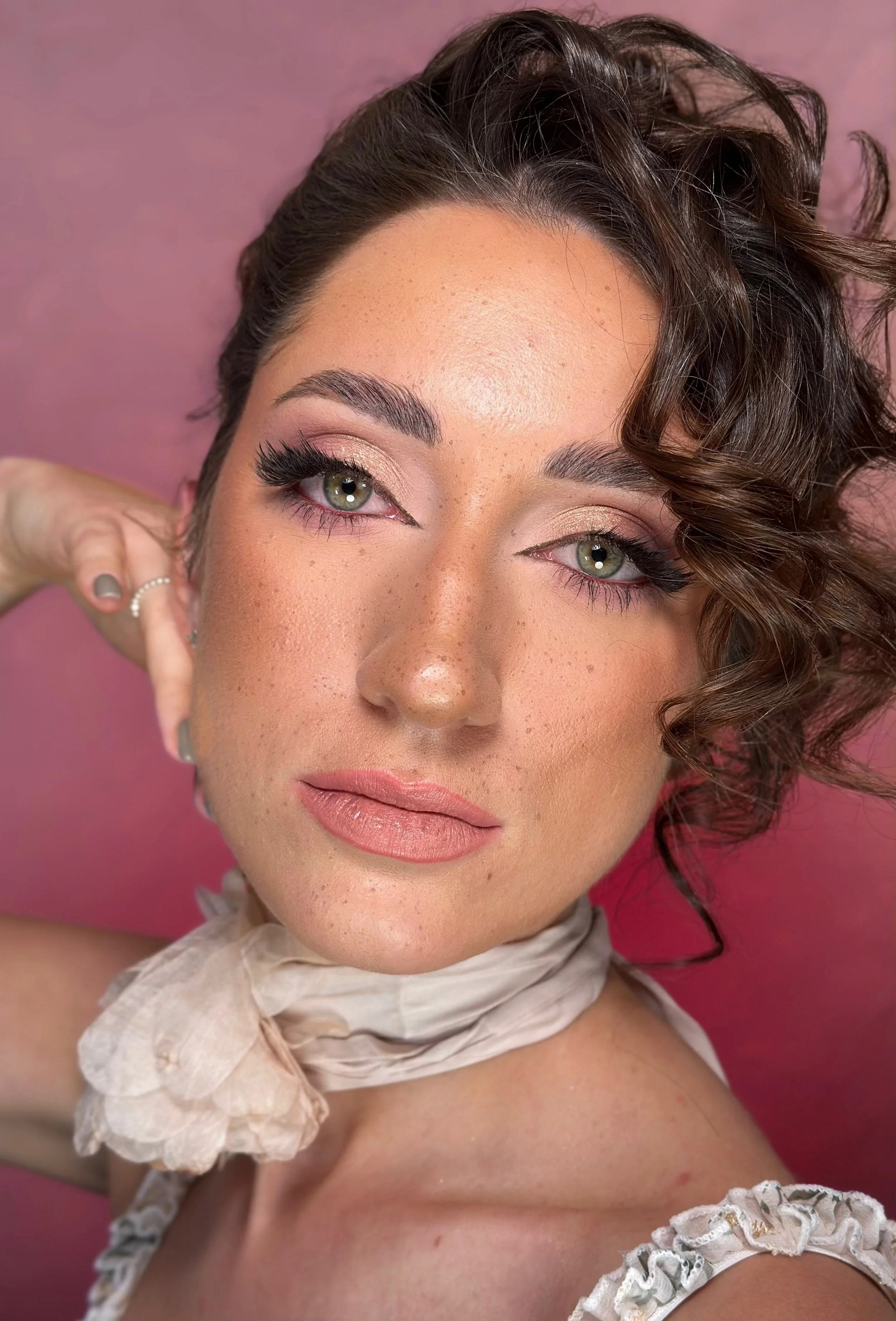 Close-up portrait of a woman with curled brown hair, wearing a soft makeup look including pink eyeshadow and nude lipstick. She has a textured scarf around her neck and a lace-trimmed outfit, against a pink background.