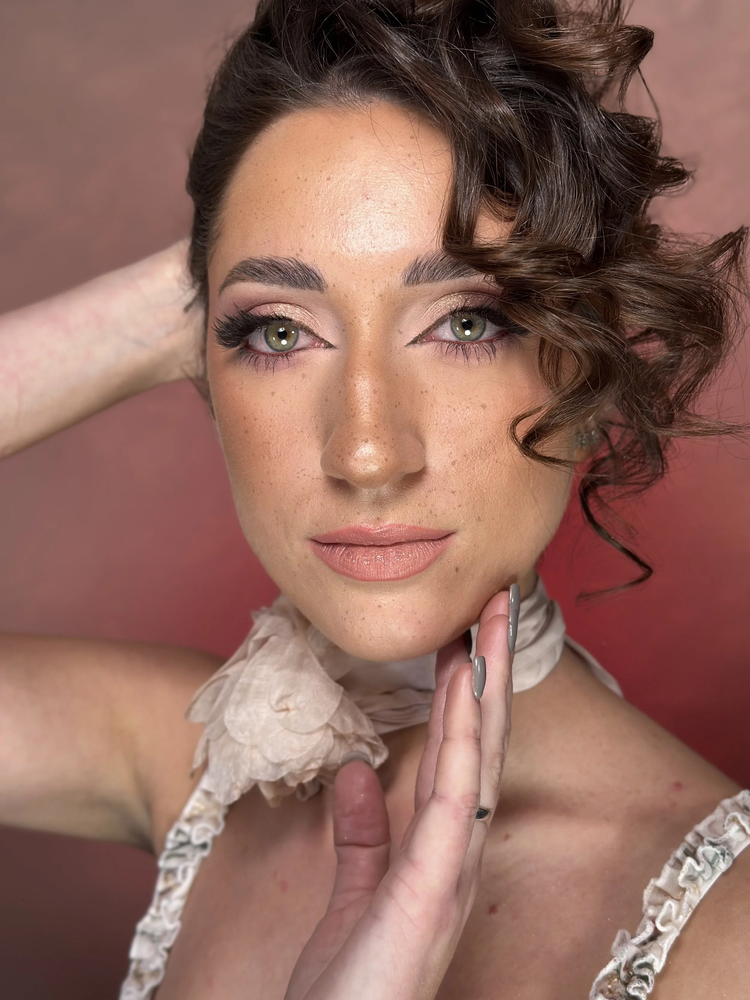 A woman with short, curly brown hair and green eyes posing with her hand near her face against a pink background.