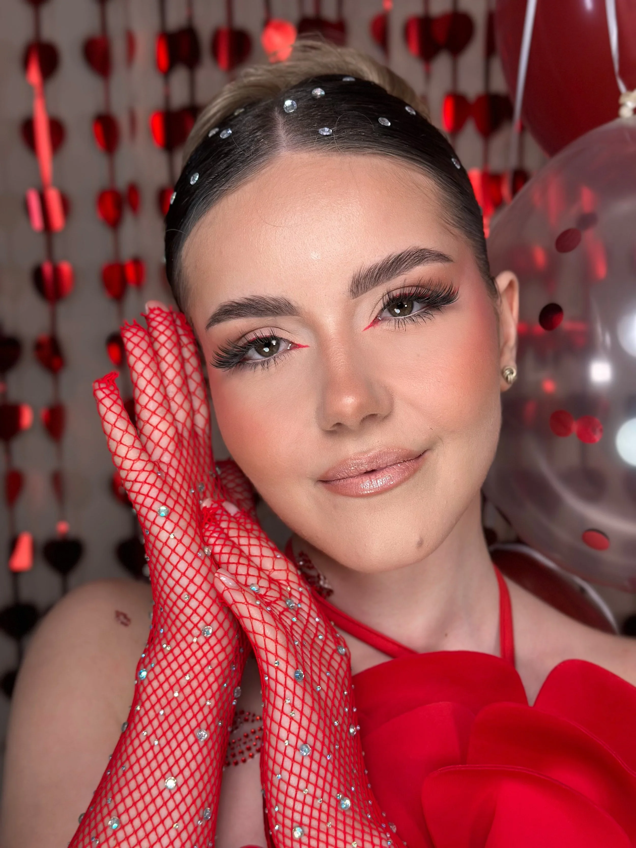 A woman wearing red mesh gloves with rhinestones, eye-catching makeup, and a red dress. She is posing in front of a backdrop decorated with red heart shapes and balloons.