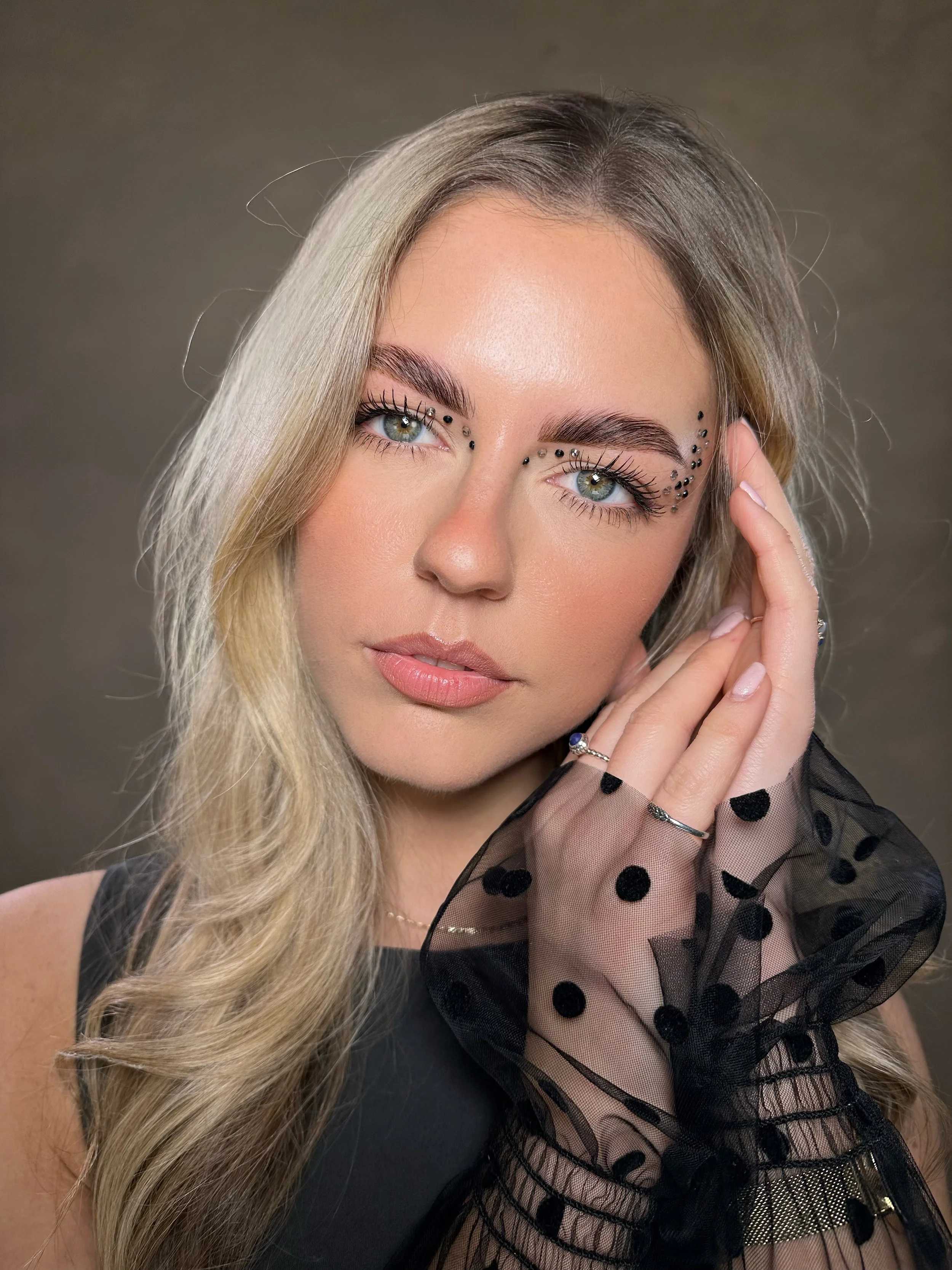 Close-up portrait of a person with long blonde hair and decorative eye makeup, wearing a black polka-dot mesh glove on one hand.