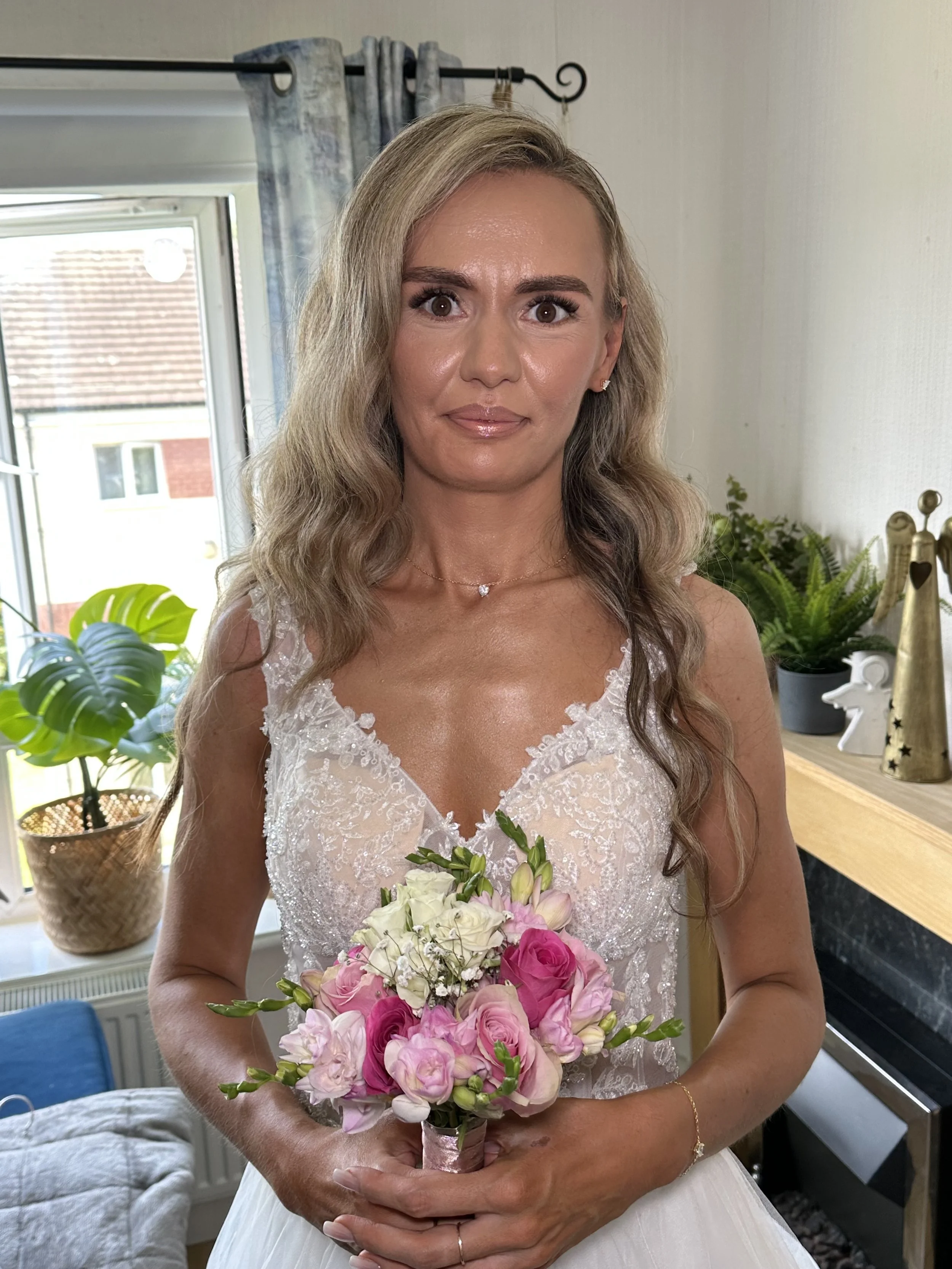 A bride with long,wavy blonde hair wearing a white lace wedding dress holding a bouquet of pink and white flowers, standing indoors near a window with plants and decorative items in the background.