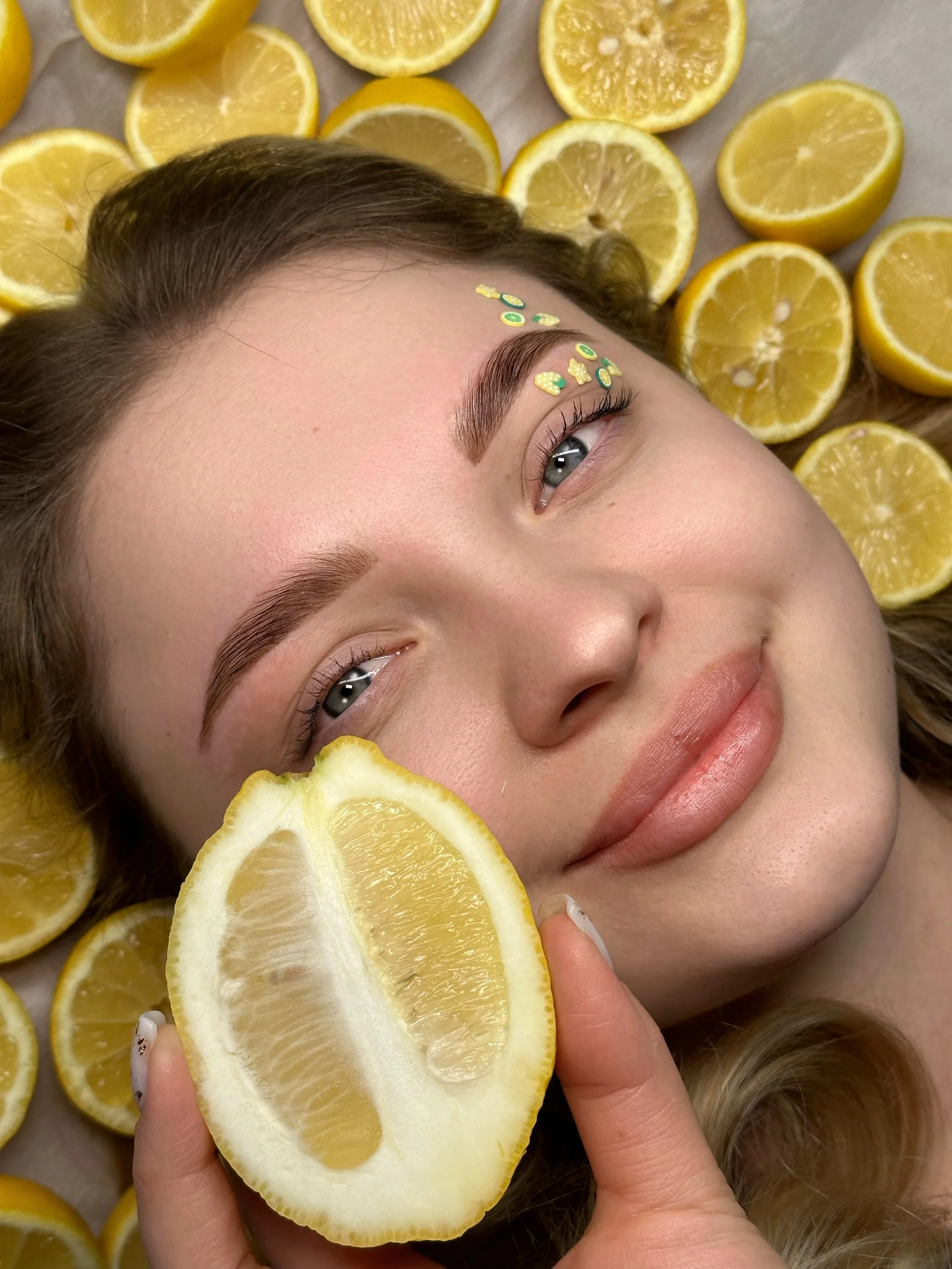 Close-up of a woman holding a lemon half, surrounded by sliced lemons, with small citrus-themed decorations above her eyebrow.