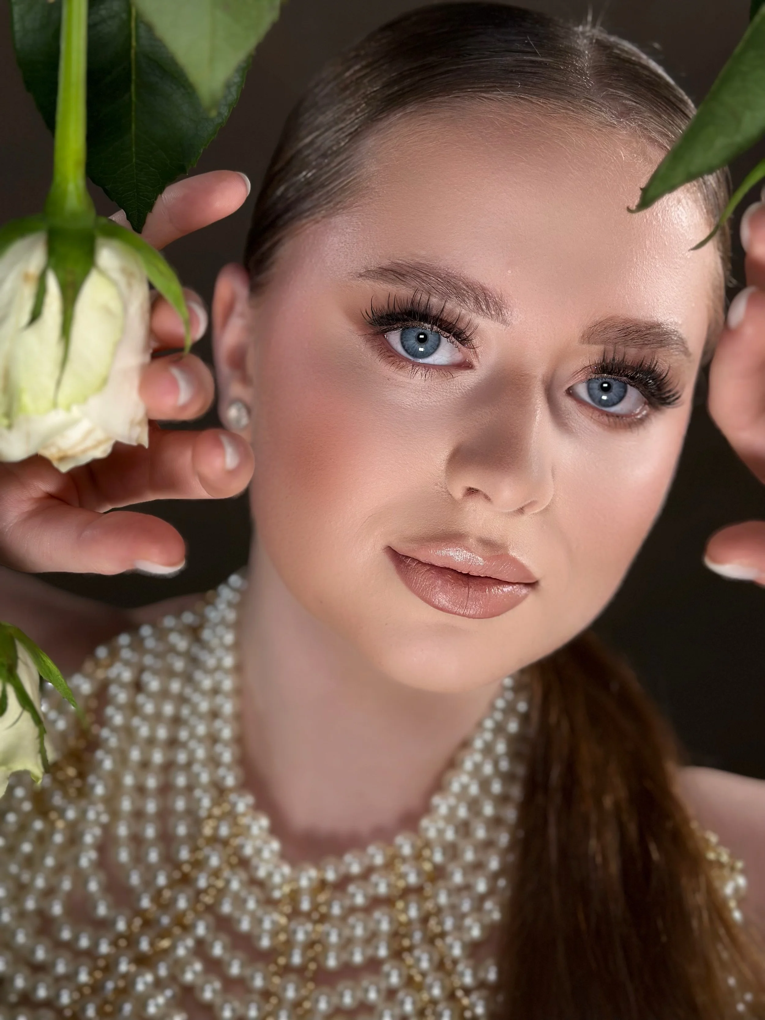 Close-up of a person with blue eyes and long lashes, wearing a pearl necklace and holding white roses, with a backdrop of green leaves.