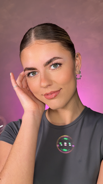 Person with slicked-back hair, wearing earrings and a gray top, posing with a colorful soap bubble in front against a pink backdrop.