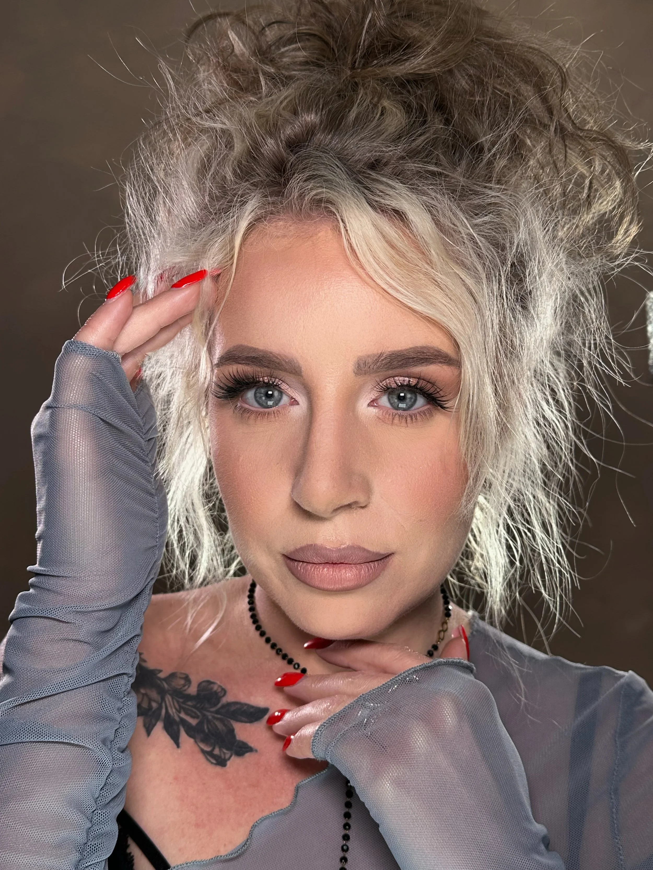 Close-up portrait of a woman with styled curly blonde hair, blue eyes, makeup, and a tattoo on her shoulder, wearing a sheer long-sleeve top and red nail polish.
