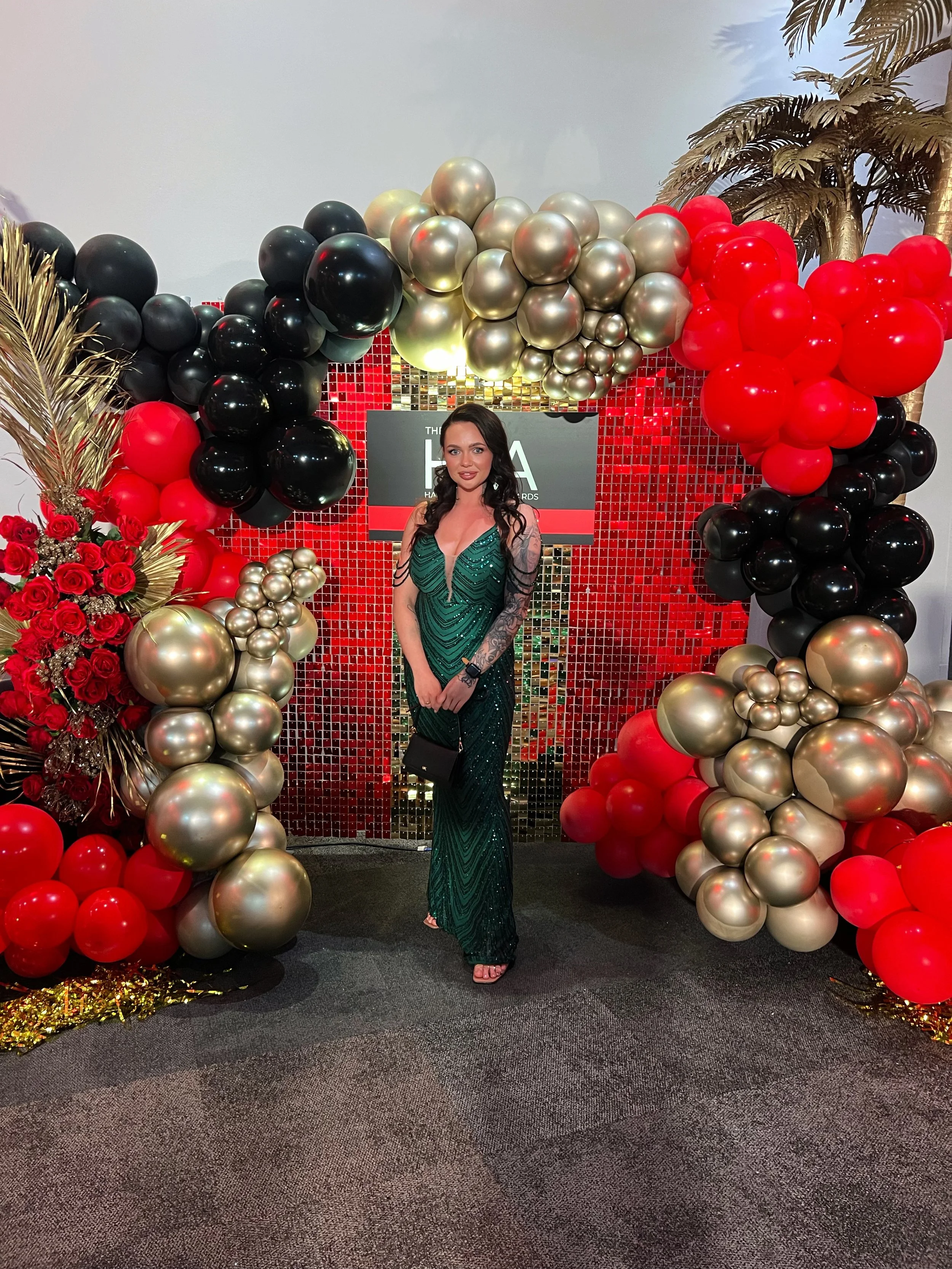 Woman in a green dress standing in front of a decorated balloon arch with black, red, and gold balloons, along with red roses and gold palm leaves.